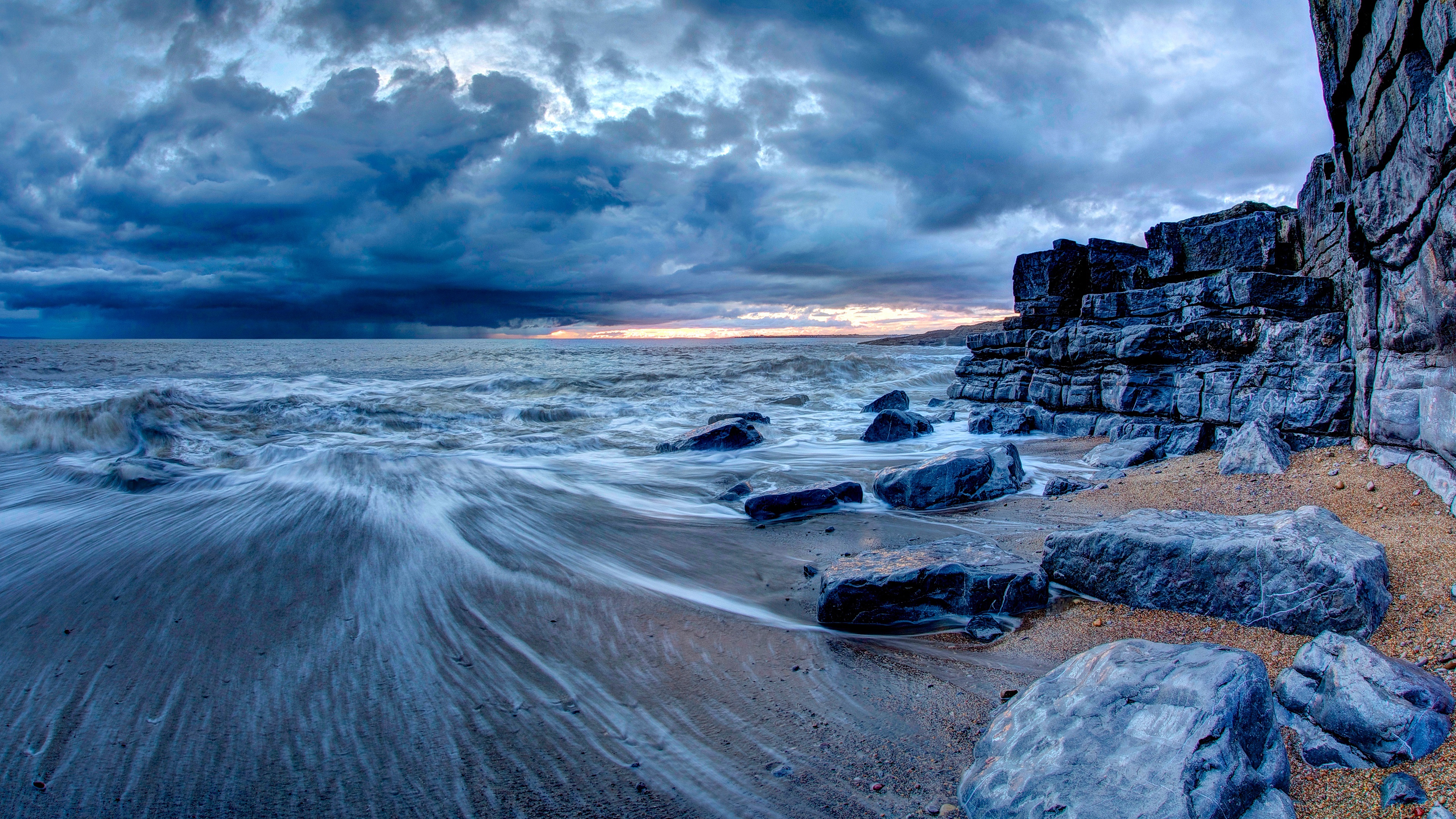 Dramatic 4K wallpaper of a rocky beach with waves crashing on the shore under a stormy sky