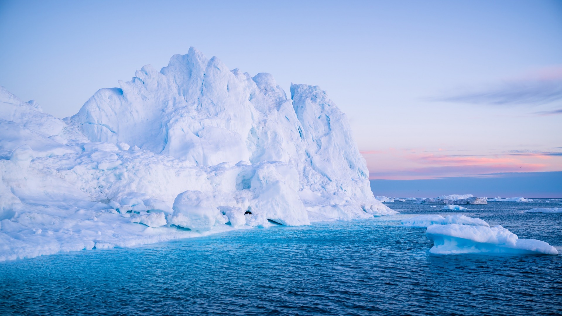 A massive iceberg towers above the tranquil blue waters, its jagged peaks catching the soft hues of a pastel sunset. Smaller ice floes dot the serene seascape, creating a breathtaking arctic panorama