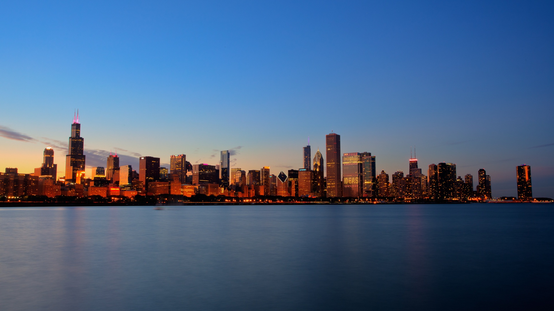 A breathtaking panorama of the Chicago skyline at dusk, with the city's iconic skyscrapers illuminated against the darkening sky and their reflections shimmering on the tranquil waters of Lake Michigan
