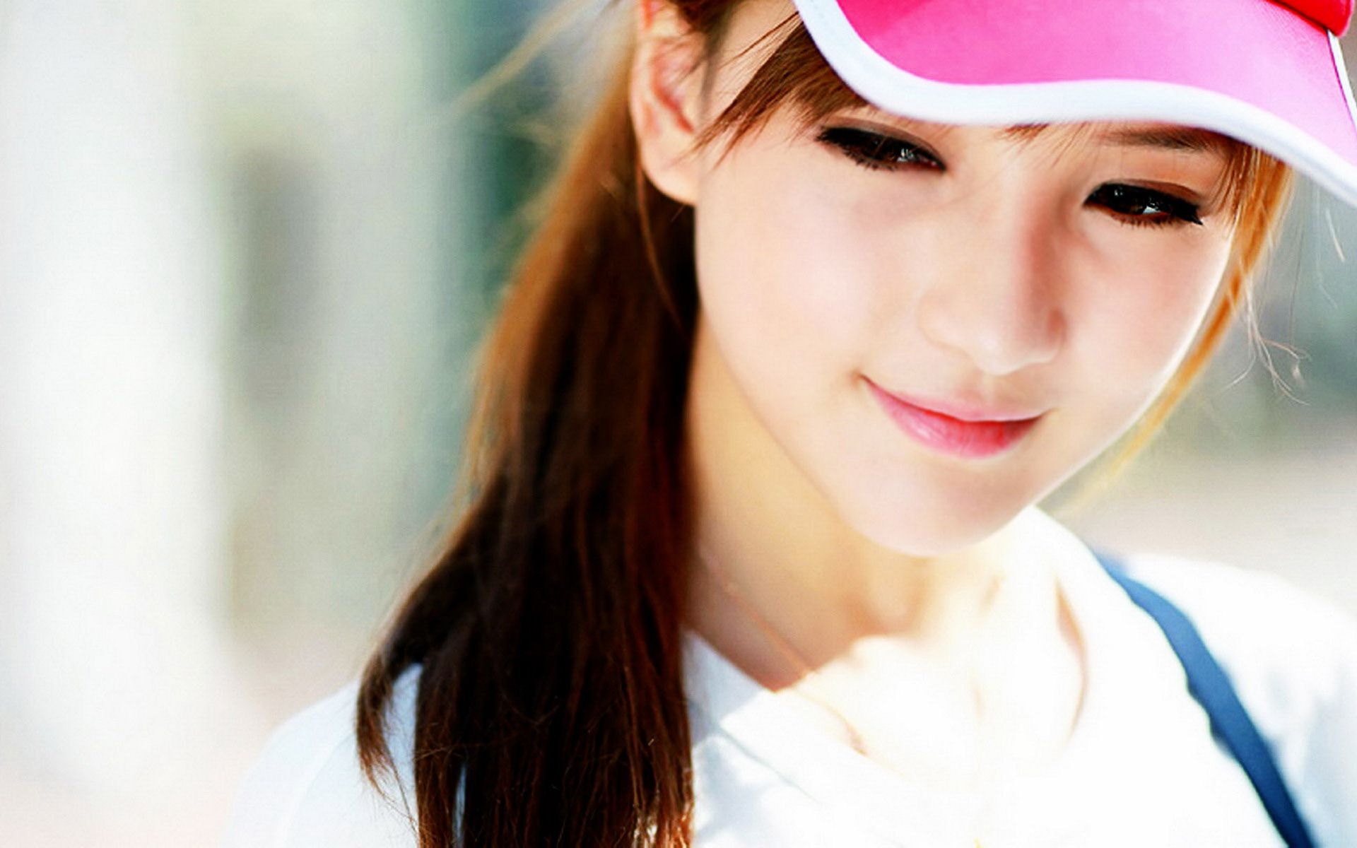 A young woman wearing a pink visor and a white shirt, looking down and smiling softly. The bright, blurred background enhances her youthful and cheerful appearance
