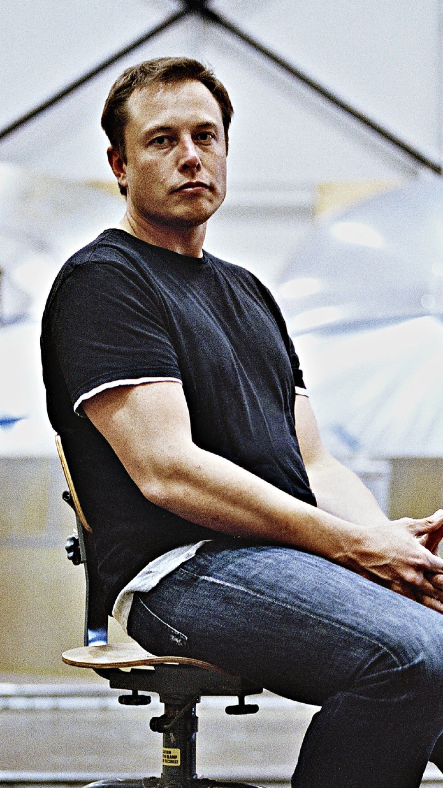 Elon Musk sitting in a chair, SpaceX factory in the background