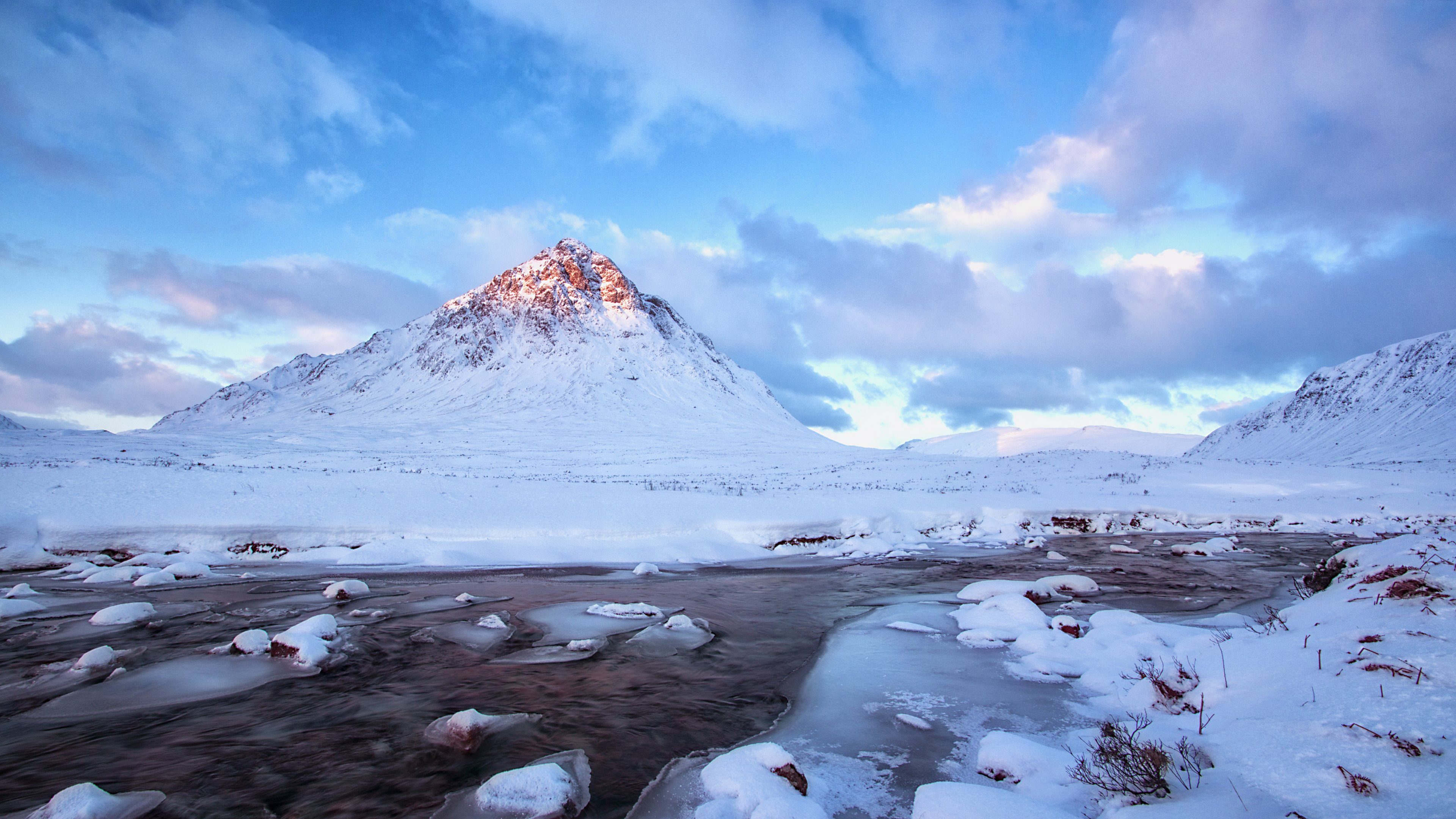 A breathtaking 4K winter landscape featuring a snow-capped mountain peak, a partially frozen river, and a vibrant sky