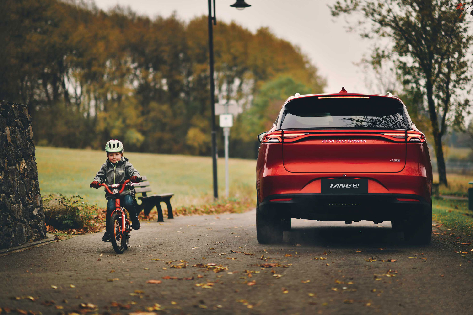 A child on a bicycle passes a red BYD Tang EV parked on a paved path in a park