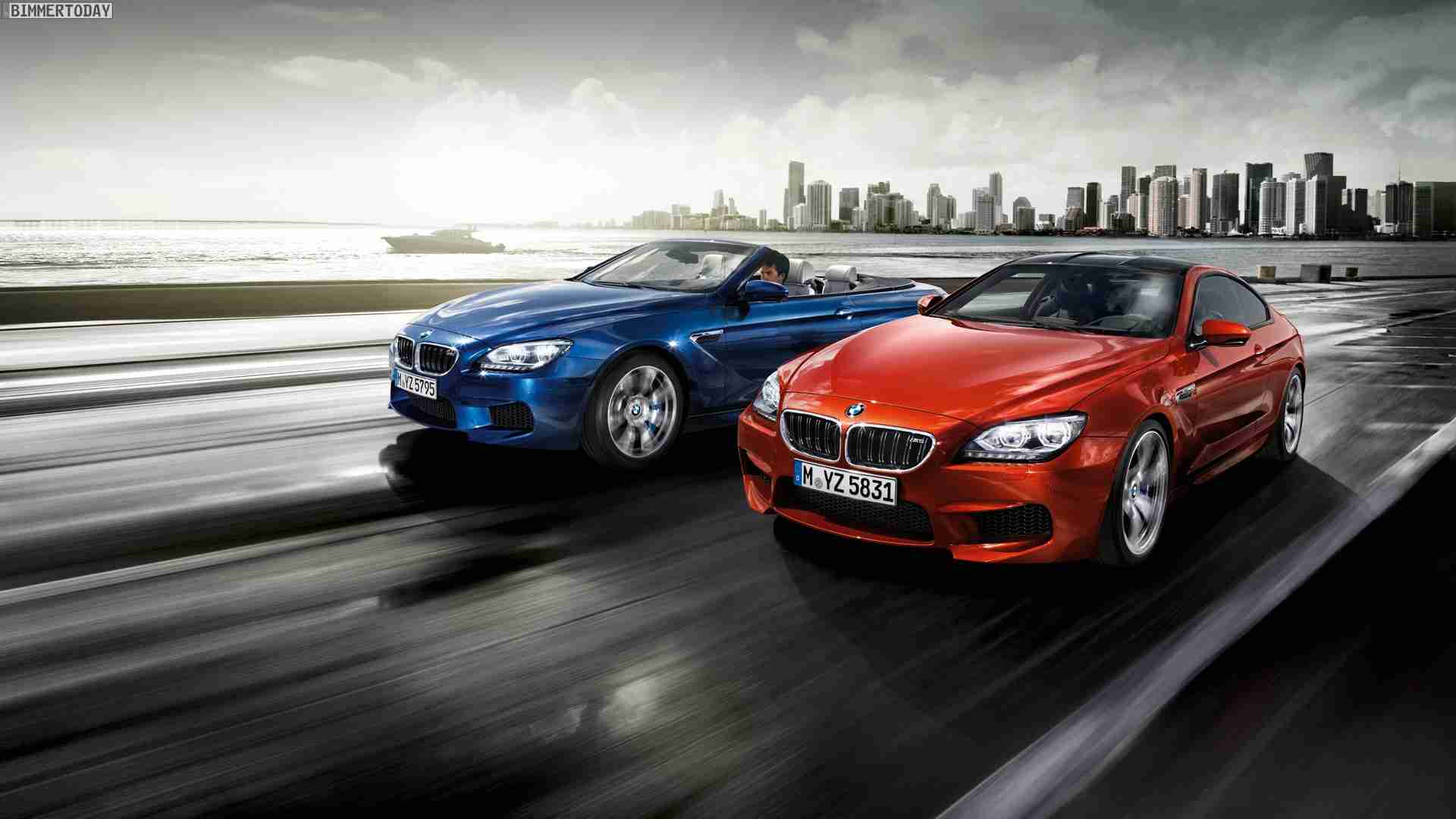 Two BMW M6 cars, a blue convertible and a red coupe, driving side by side on a coastal highway with a cityscape in the background