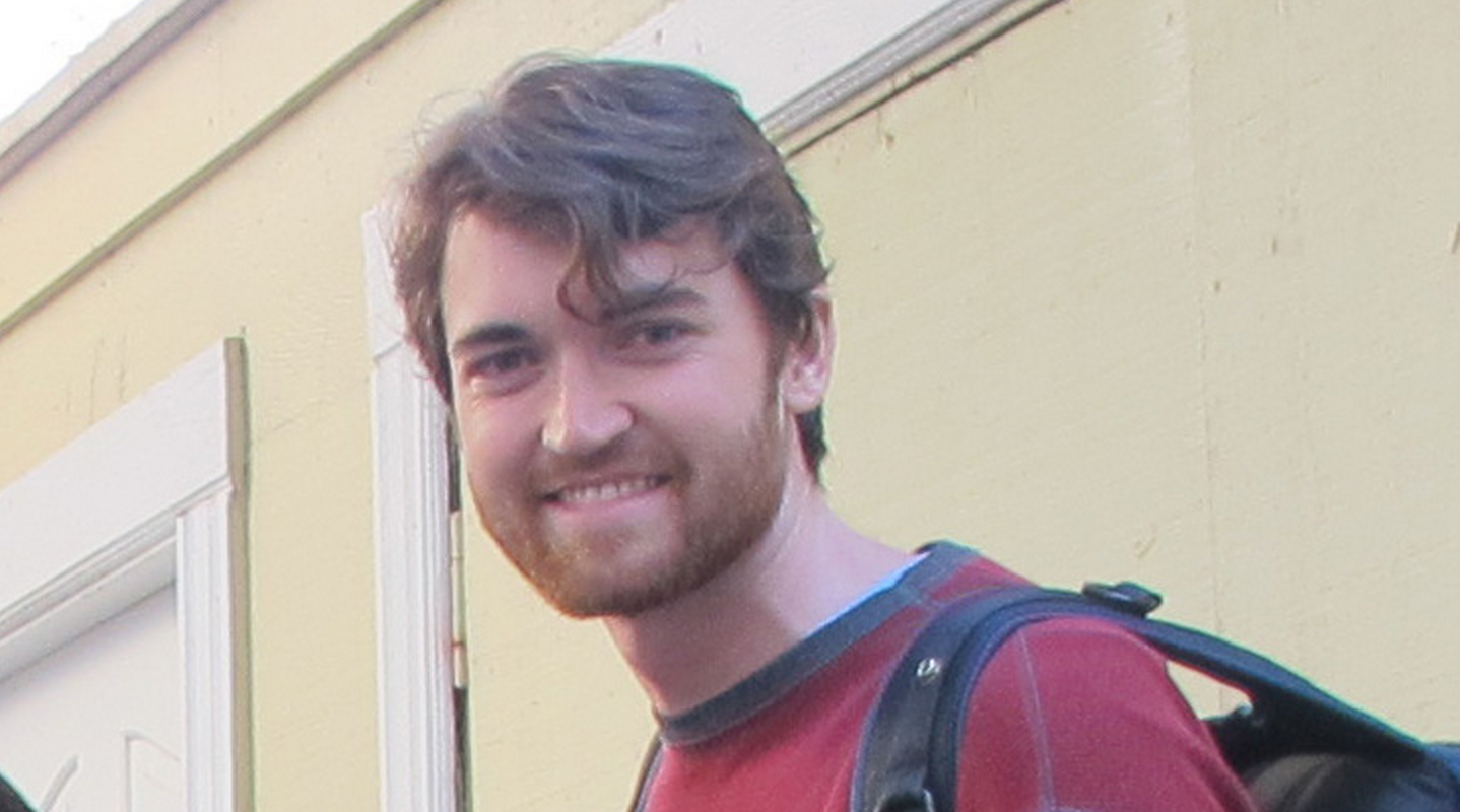 A young man with light brown hair and a beard, wearing a red shirt and backpack, smiles at the camera