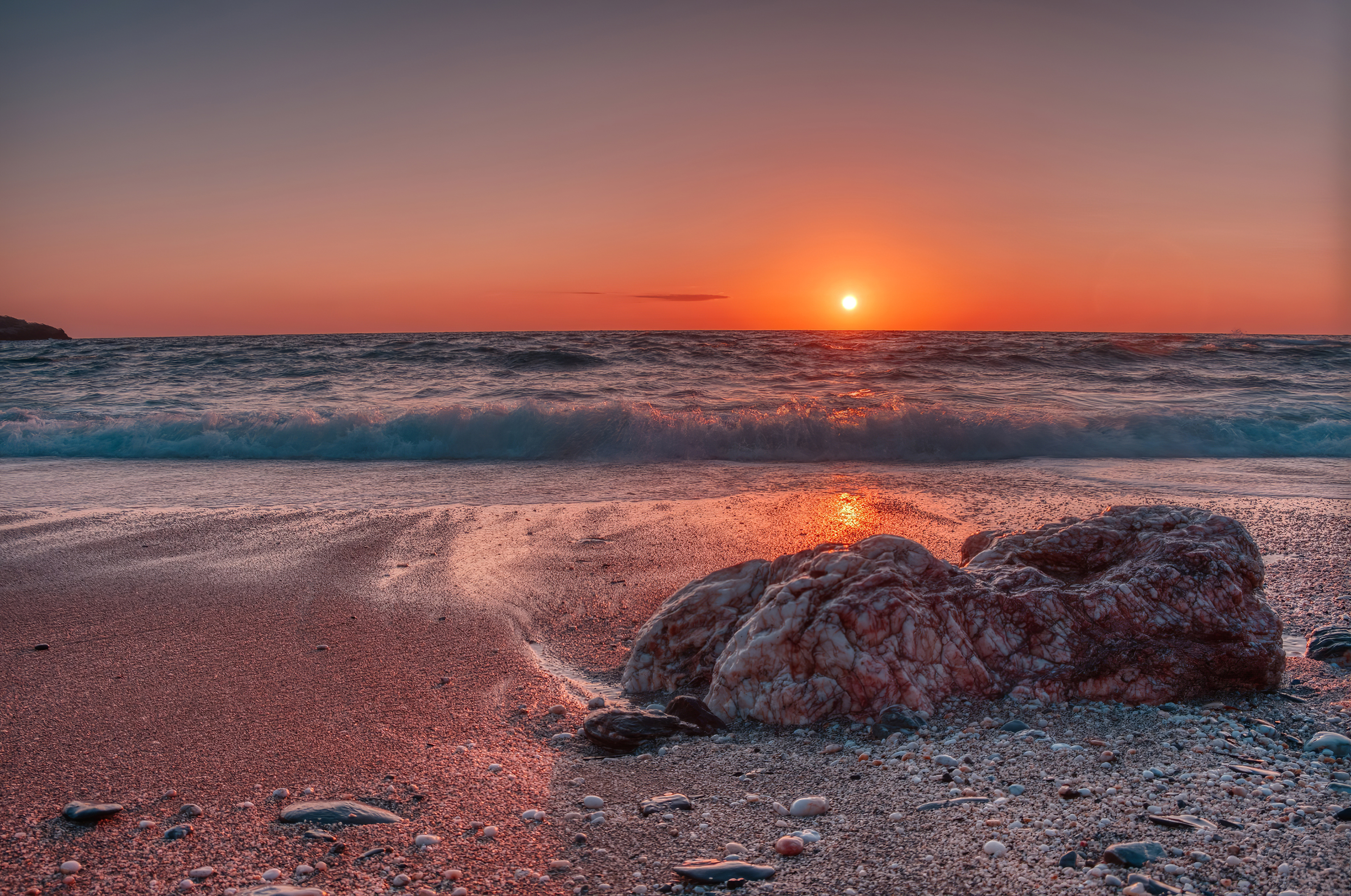 A breathtaking 4k wallpaper of a vibrant sunset over a tranquil beach. The warm hues of the setting sun paint the sky in shades of orange and pink, reflecting on the gentle waves lapping at the sandy shore. A large, textured rock rests on the beach, adding a touch of natural beauty to the scene