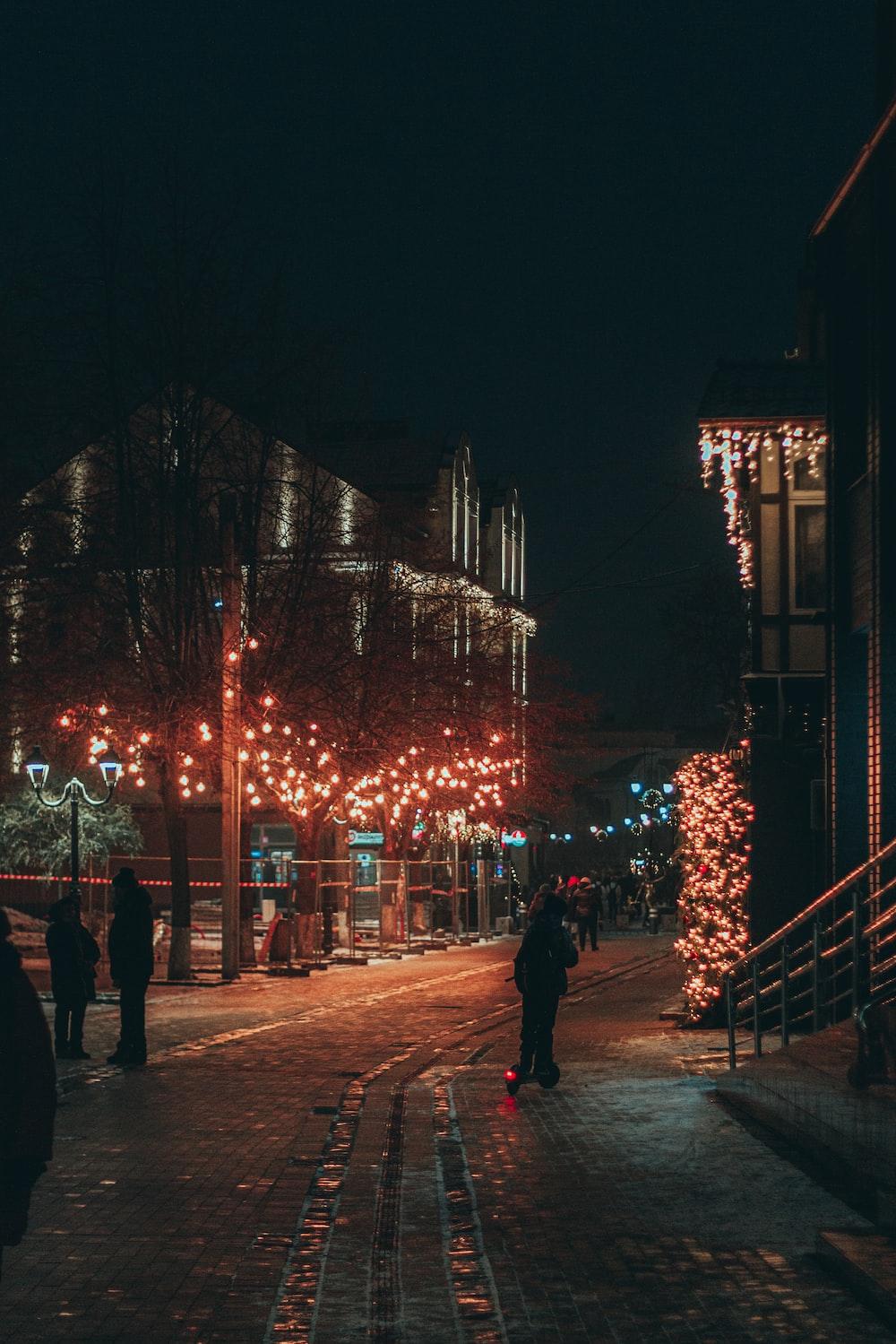 A Group Of People Walking On Sidewalk With Christmas Lights