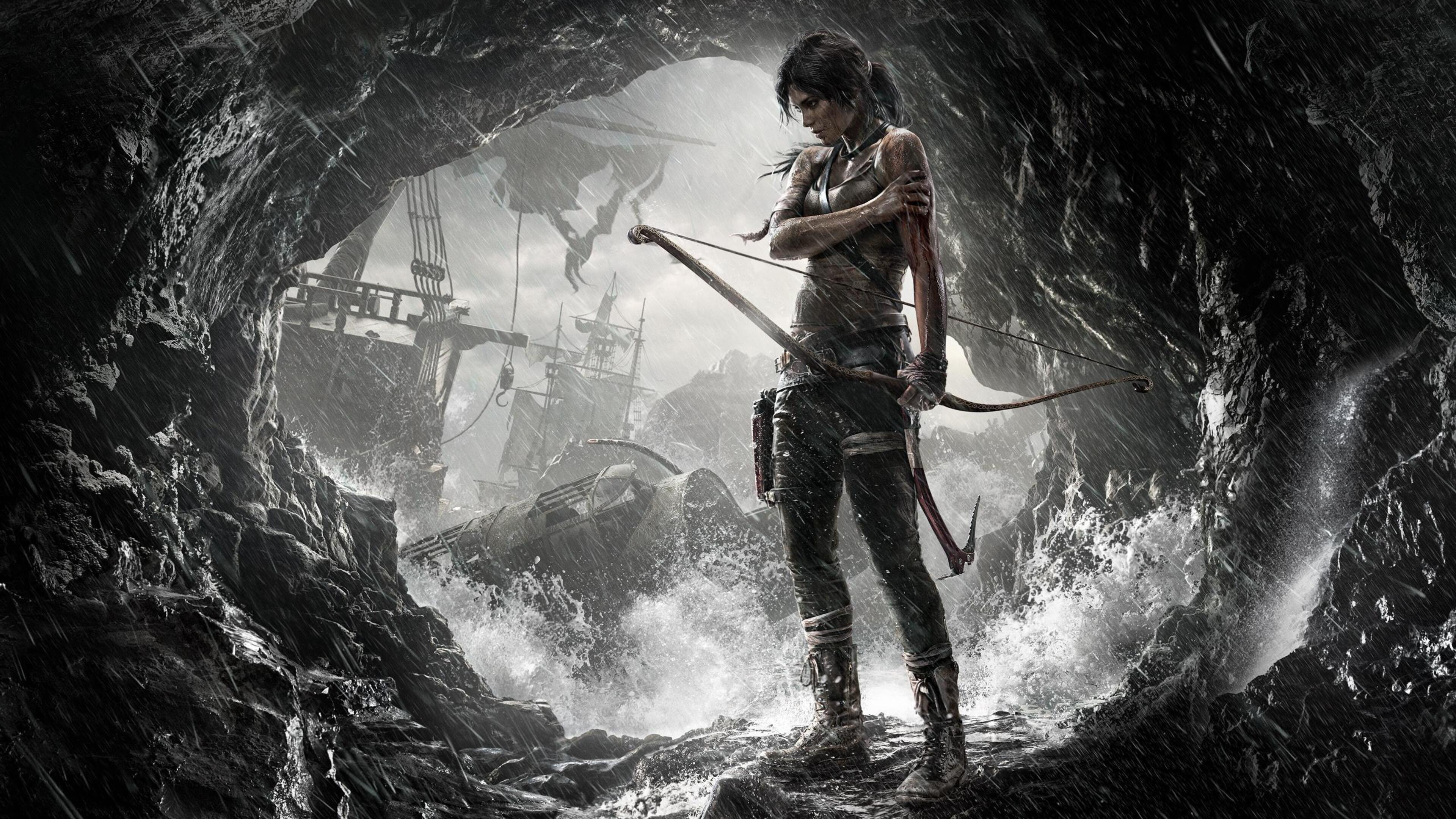 Lara Croft stands defiantly in a stormy sea cave, bow in hand, with a wrecked ship in the background, in this dramatic wallpaper from the Tomb Raider reboot