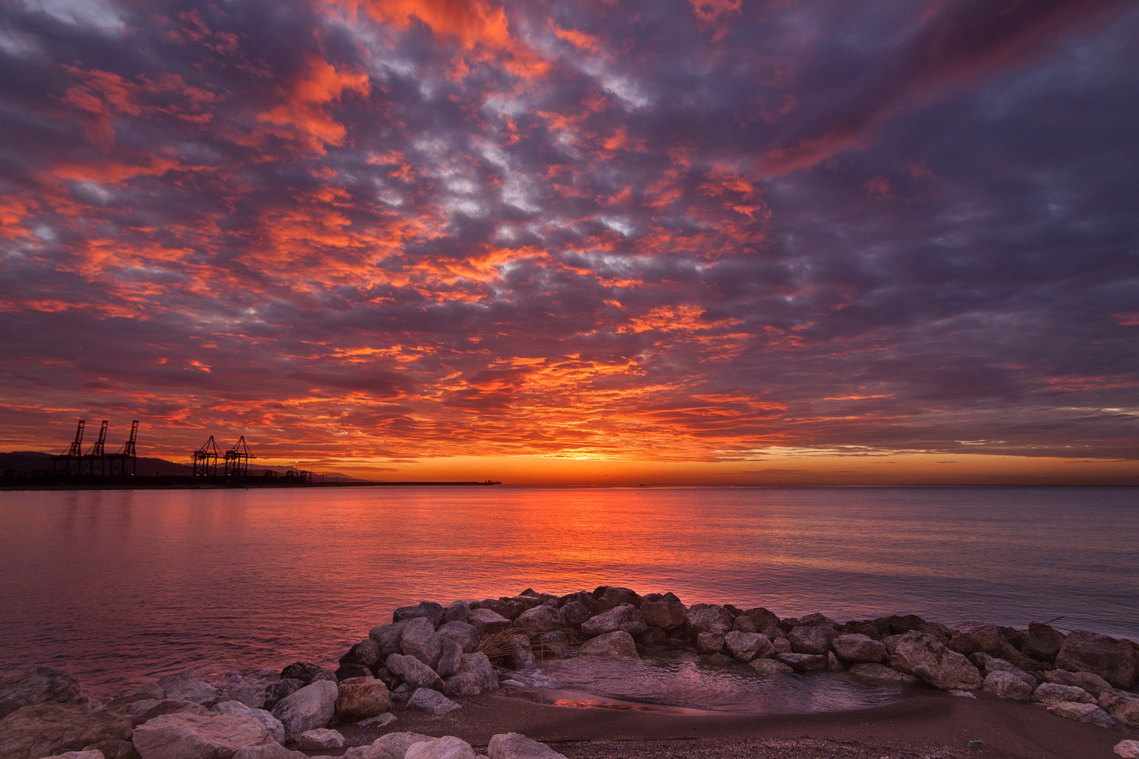 A breathtaking 4K wallpaper capturing a vibrant sunrise over a calm sea, with the silhouette of industrial cranes in the distance and a rocky shoreline in the foreground
