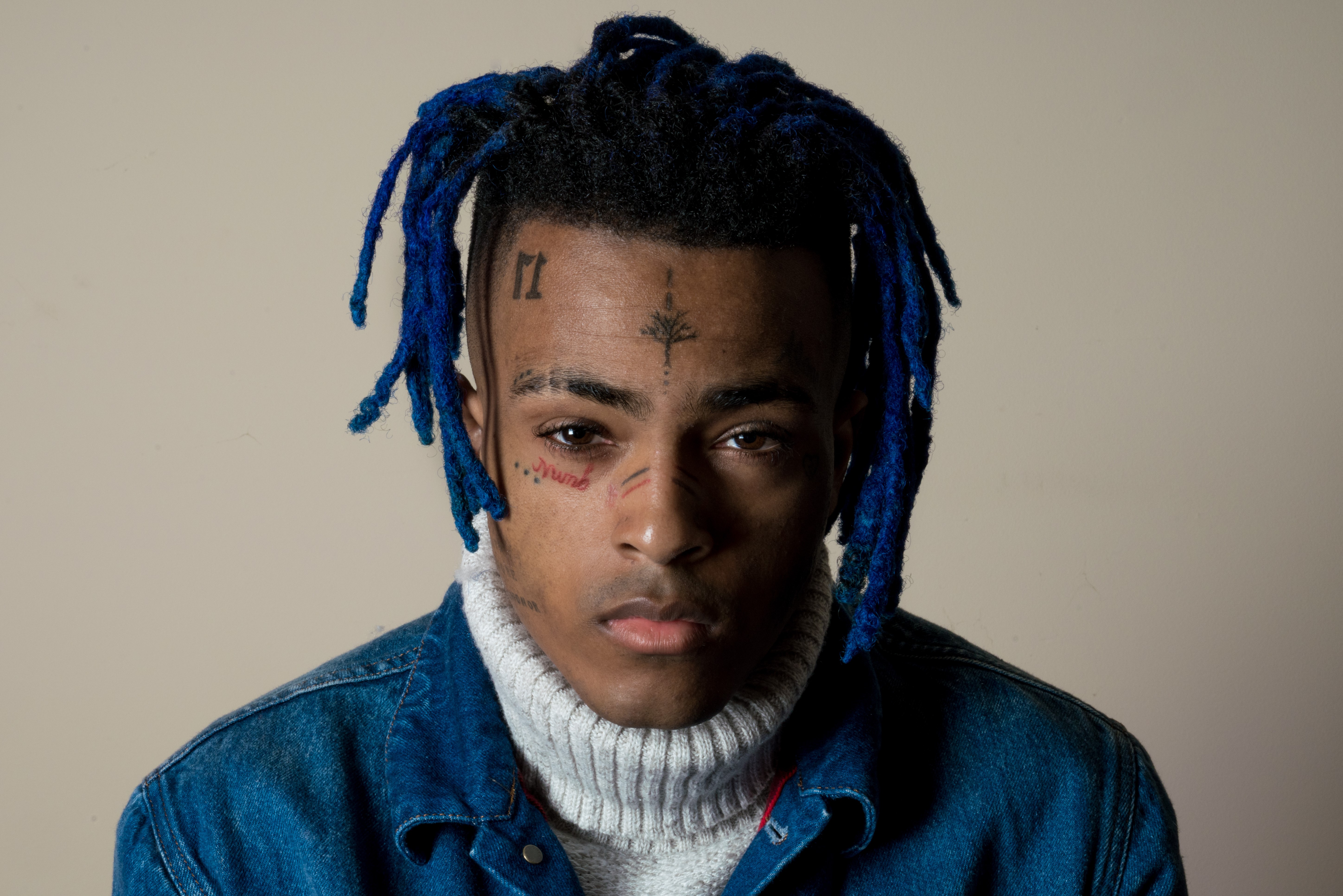 Close-up portrait of XXXTentacion with blue dreadlocks, wearing a denim jacket and a grey turtleneck