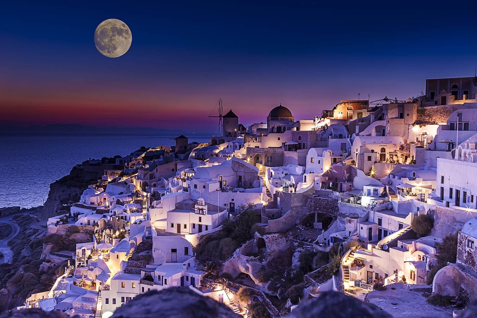 A breathtaking night view of Oia, Santorini, Greece, with white-washed buildings cascading down the cliffs, illuminated by the soft glow of lights under a full moon