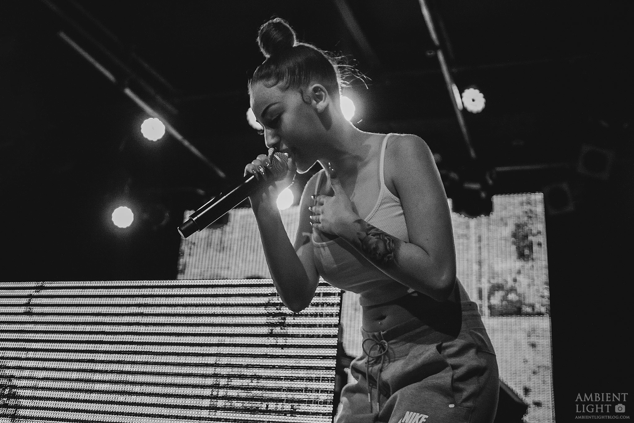 Bhad Bhabie performing live on stage, holding a microphone and singing. She's wearing a white tank top and grey sweatpants