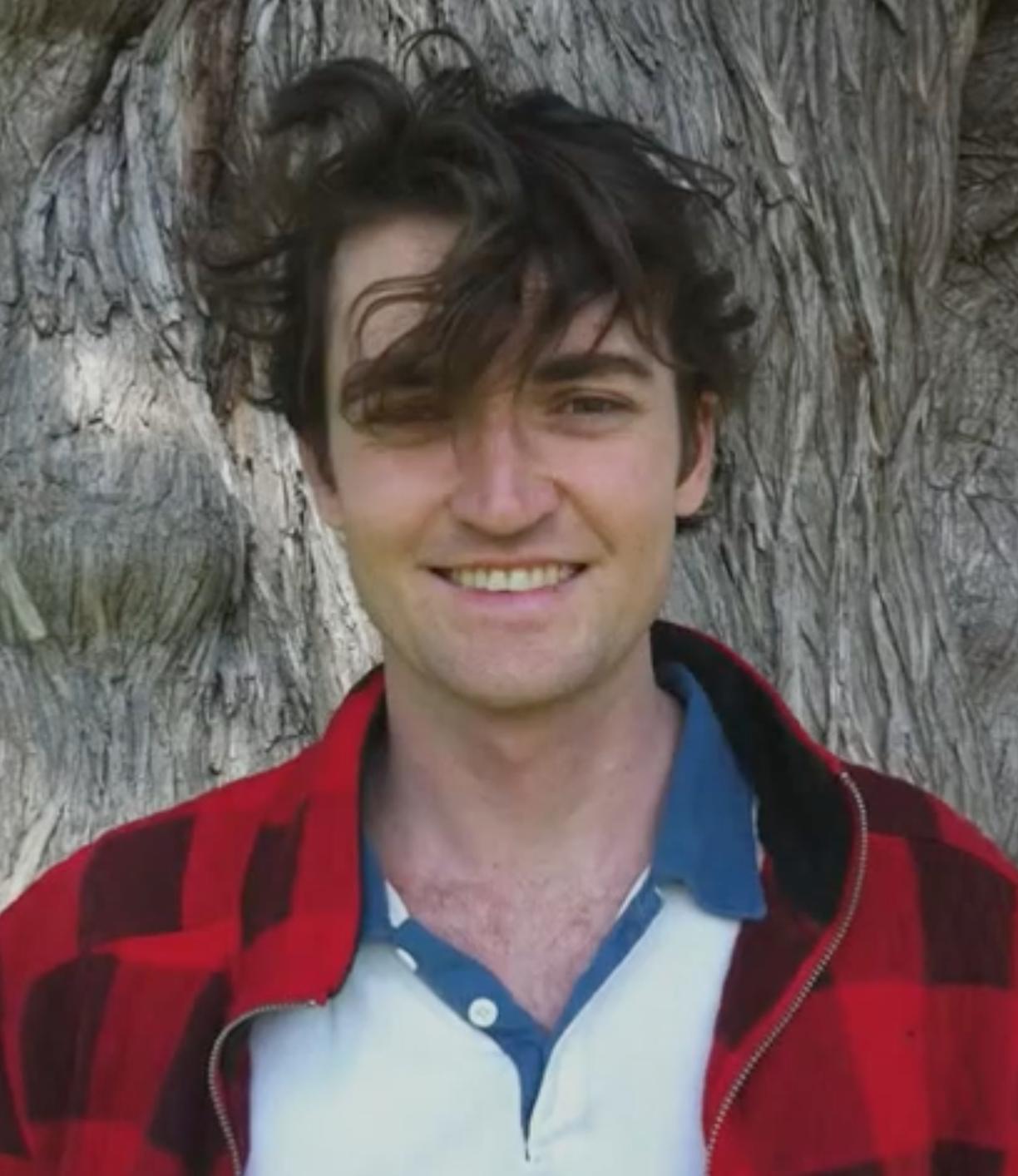 A young man with messy brown hair, wearing a red and black plaid jacket and a white polo shirt, stands in front of a tree