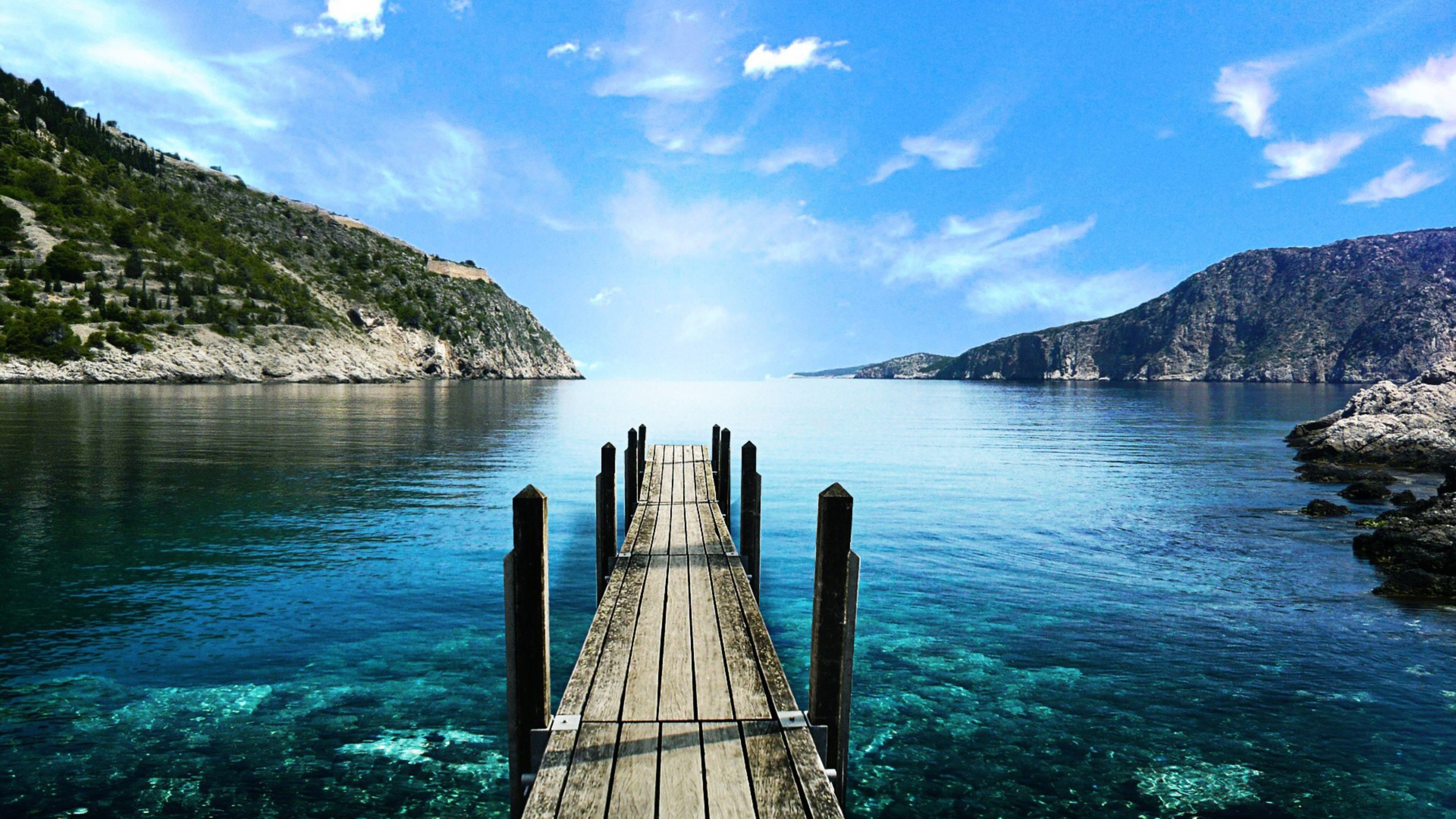 A serene 4K wallpaper featuring a wooden pier extending into crystal-clear turquoise water, nestled between lush green hills under a vibrant blue sky