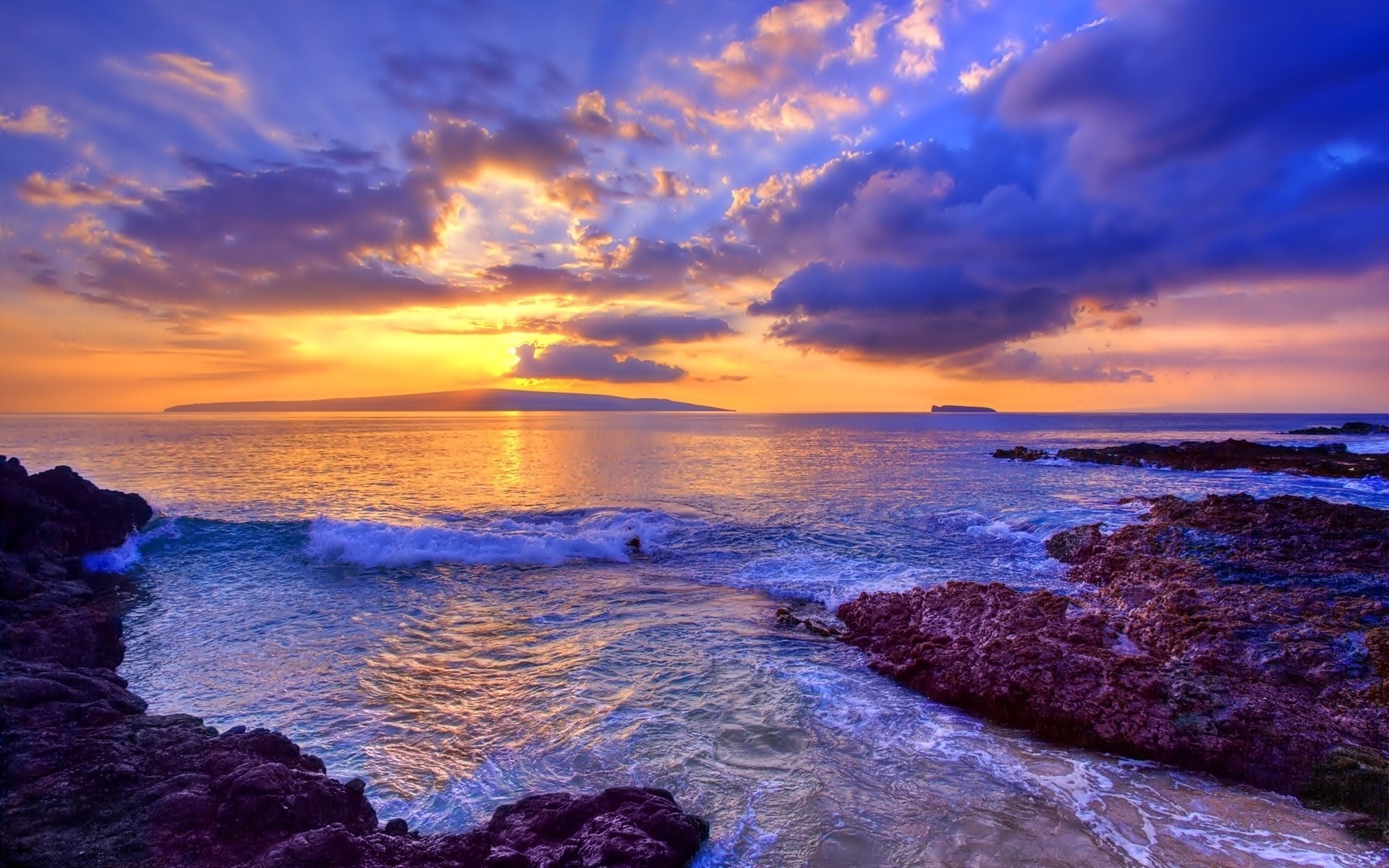 A breathtaking sunset over the ocean with vibrant purple and orange hues reflecting on the water's surface. The foreground showcases dark volcanic rocks, while gentle waves create a tranquil scene