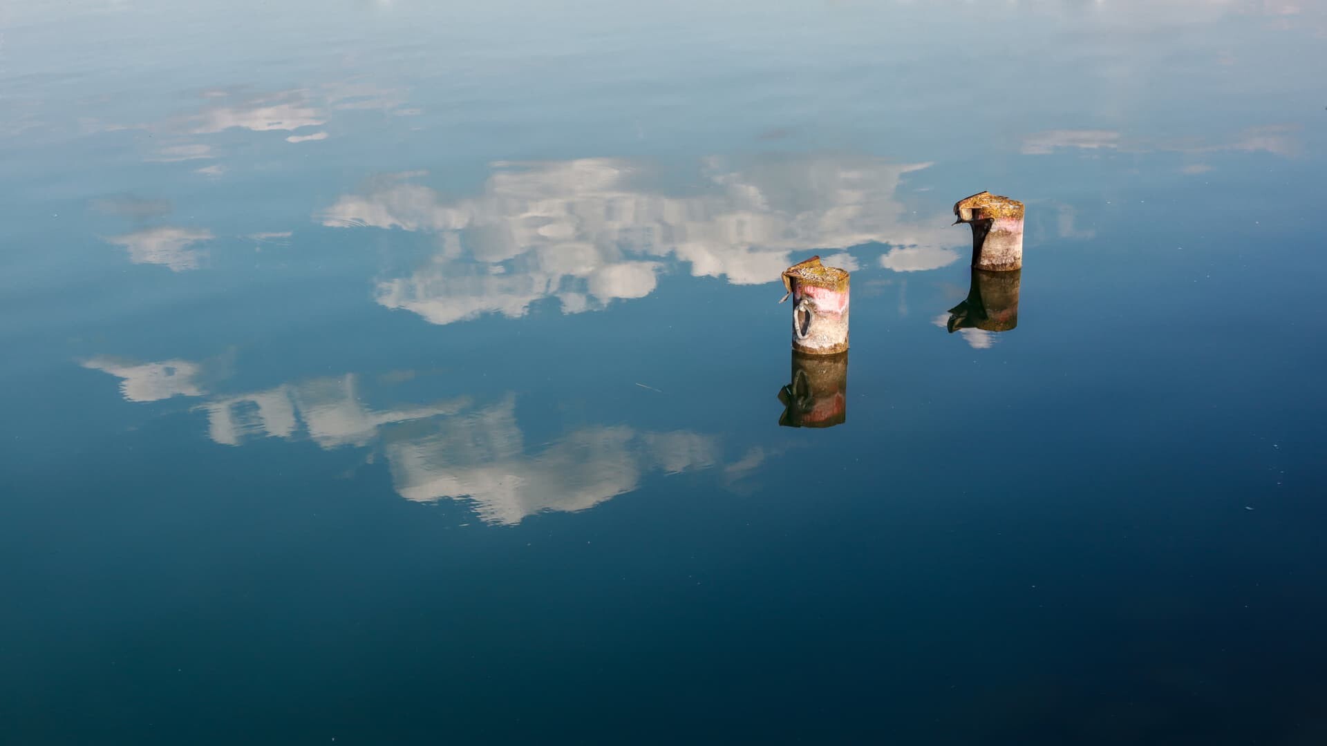 Two weathered mooring posts reflected in the calm, blue water of a lake or harbor, with a soft reflection of clouds in the background