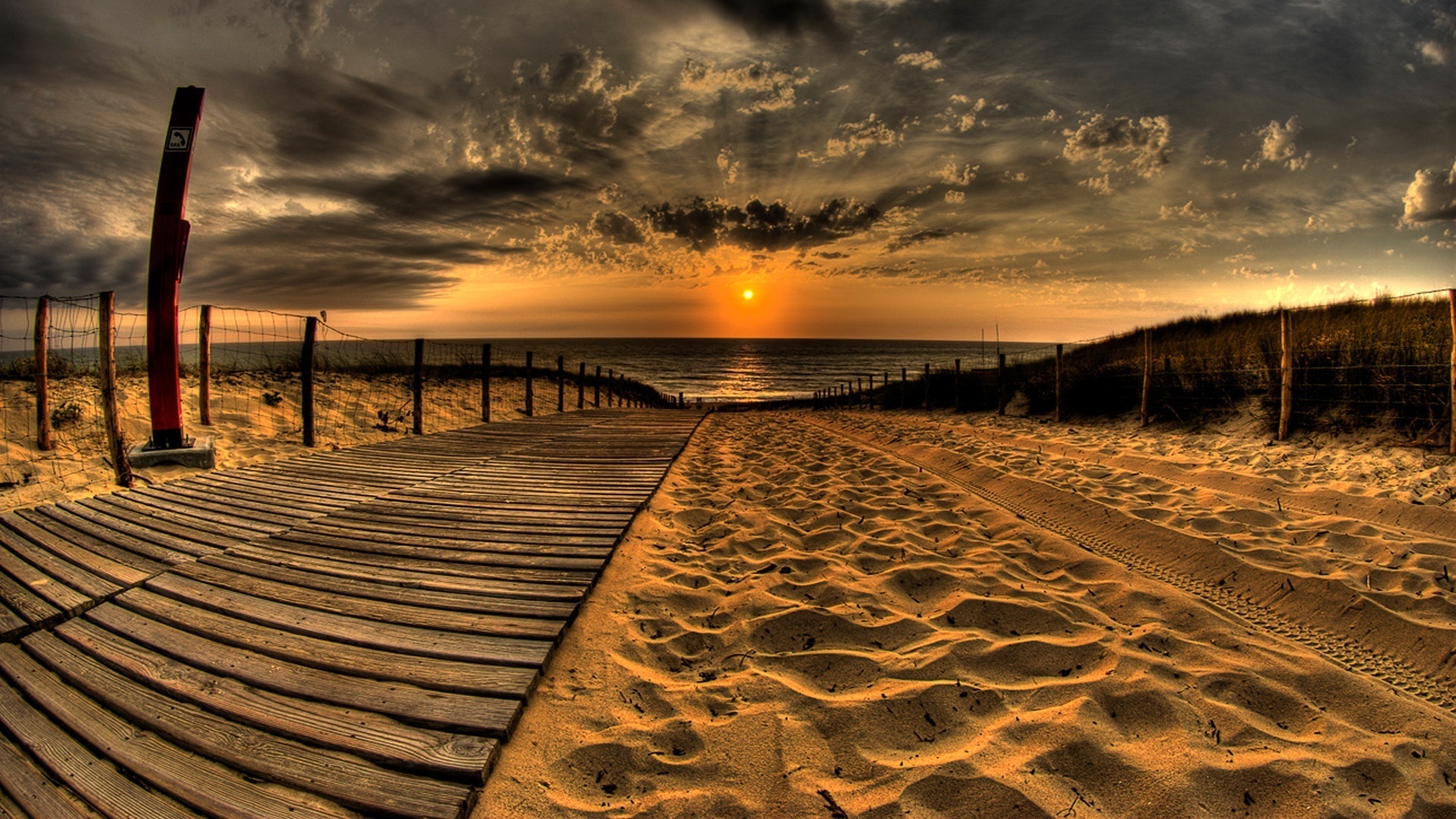 A wooden boardwalk leads towards the ocean at sunset on a sandy beach. Tire tracks are visible in the sand