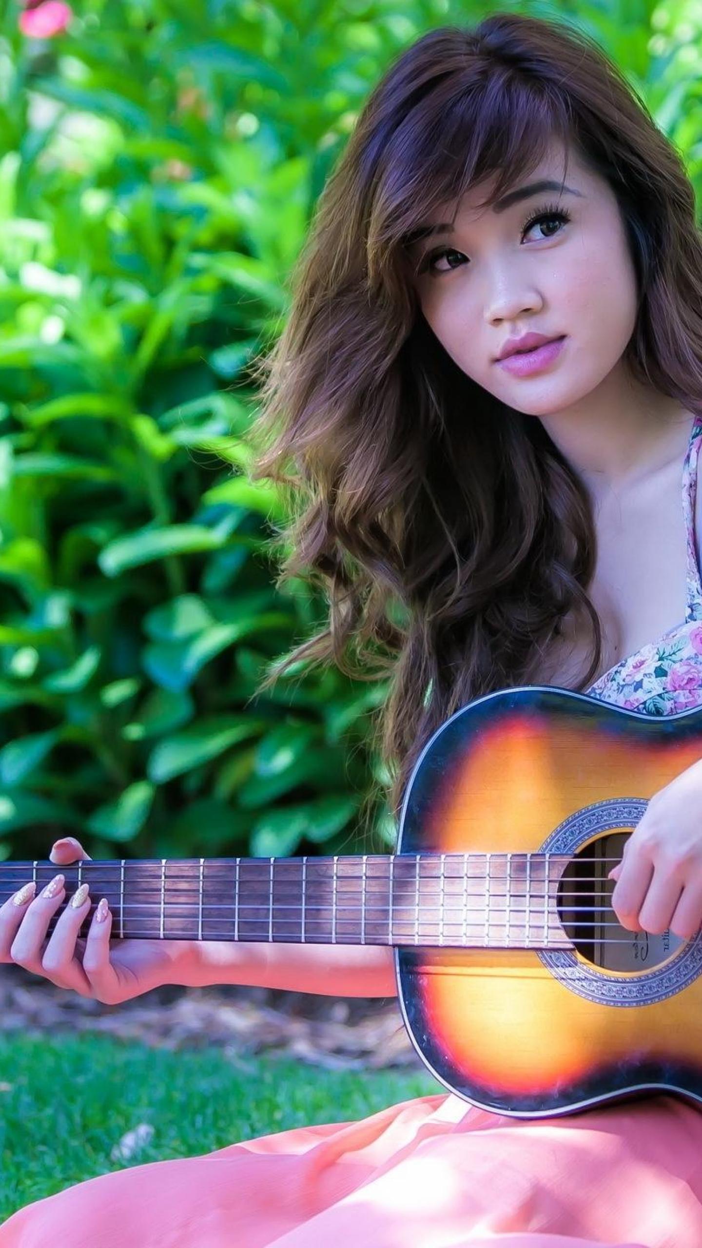 A young woman sits outdoors with her acoustic guitar, looking serenely at the camera