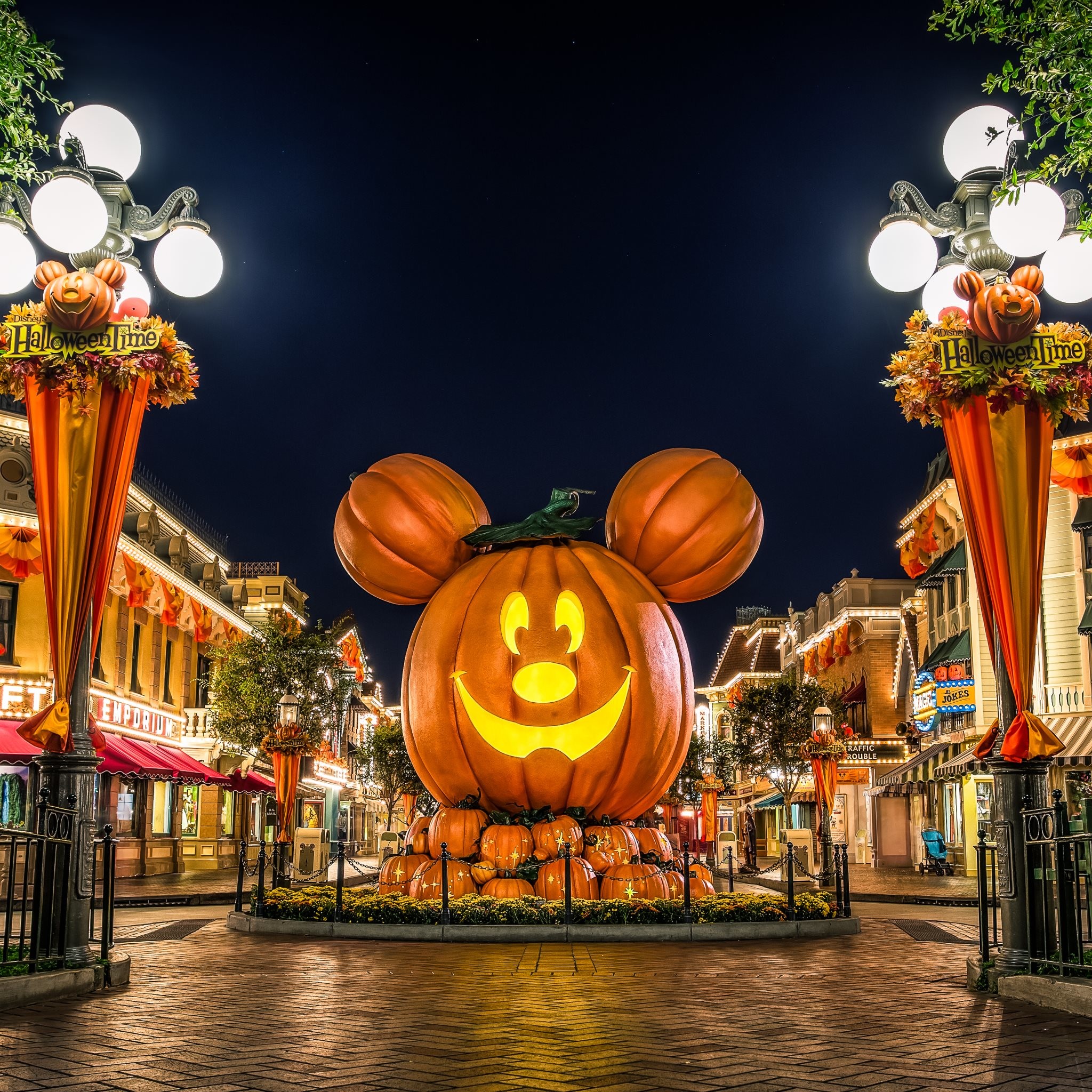A giant Mickey Mouse-shaped pumpkin illuminates a Disney theme park street at night, decorated for Halloween