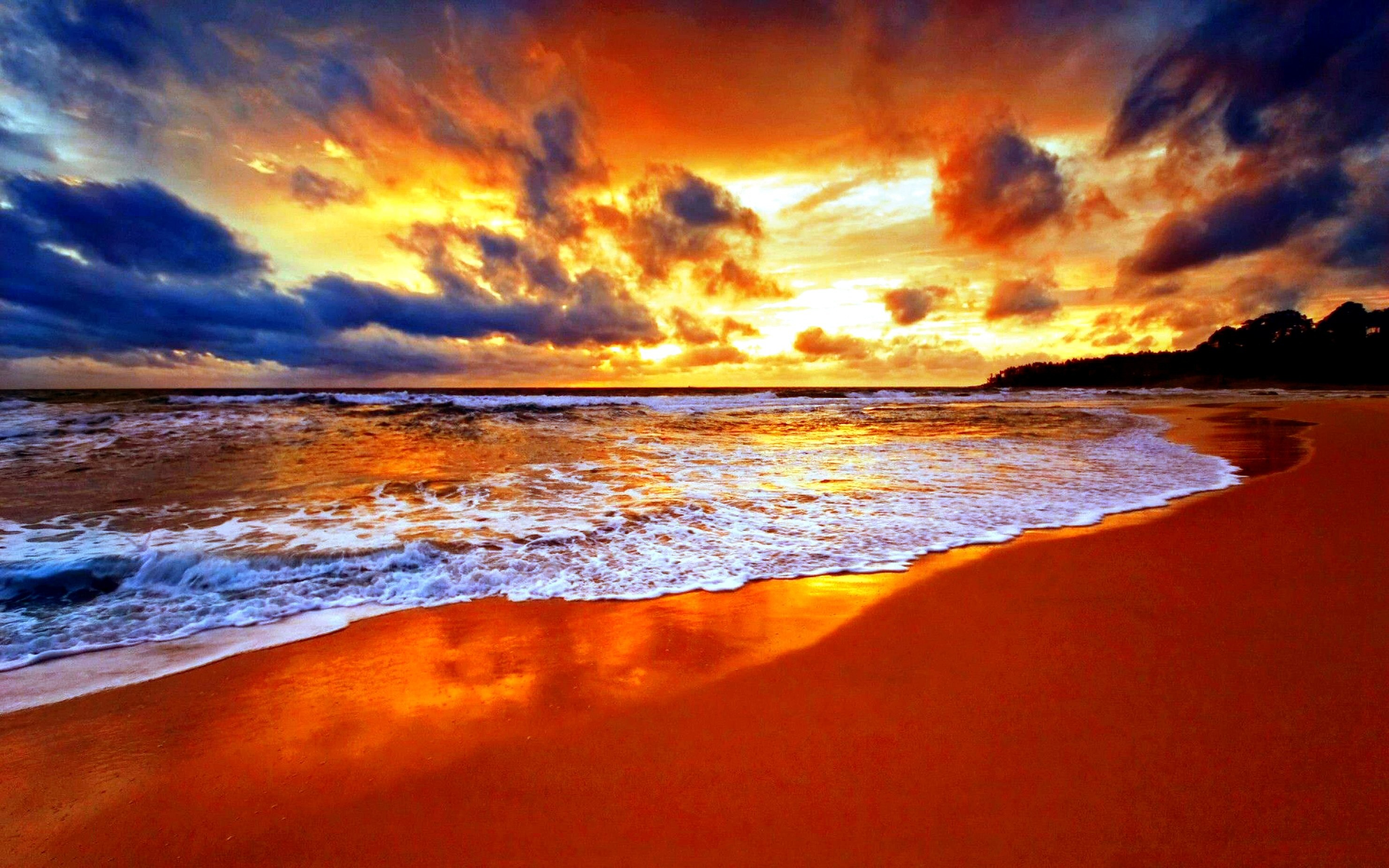 A breathtaking sunset over a vibrant beach, with fiery orange and gold hues reflecting on the wet sand and gentle waves. The dramatic sky blends shades of blue, purple, and orange, creating a stunning natural wallpaper