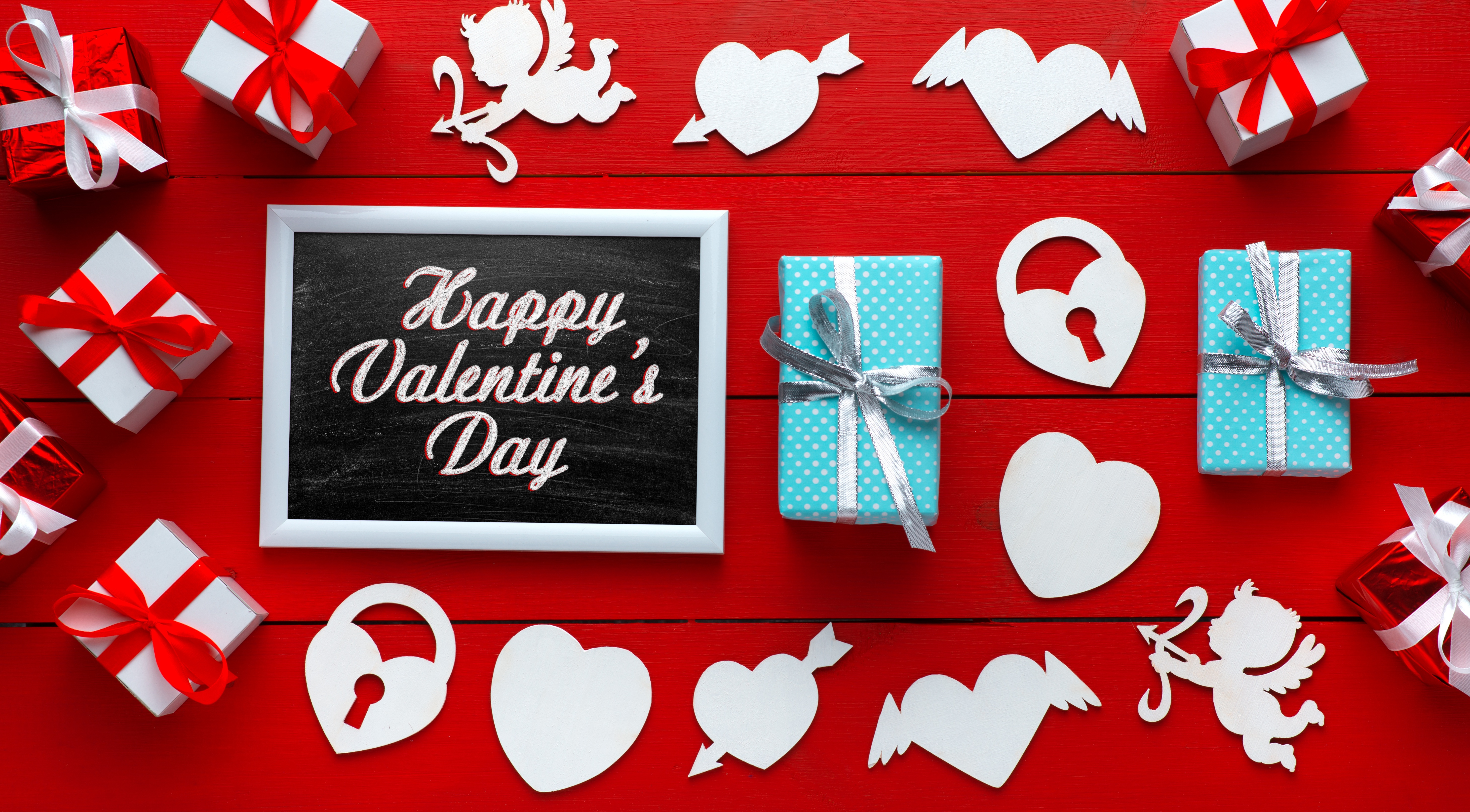 A vibrant flat lay for Valentine's Day with gifts, hearts, cupids, and a chalkboard message on a red wooden background