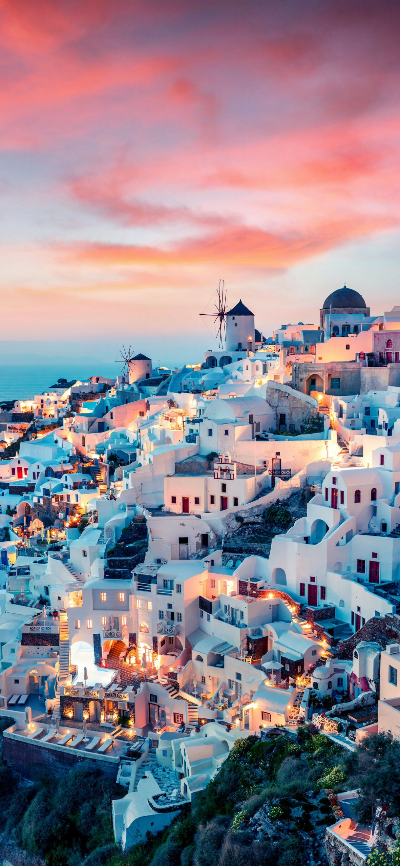 White-walled buildings perched on a cliffside in Santorini, Greece, overlooking the Aegean Sea at sunset. Windmills stand tall against the vibrant hues of the sky
