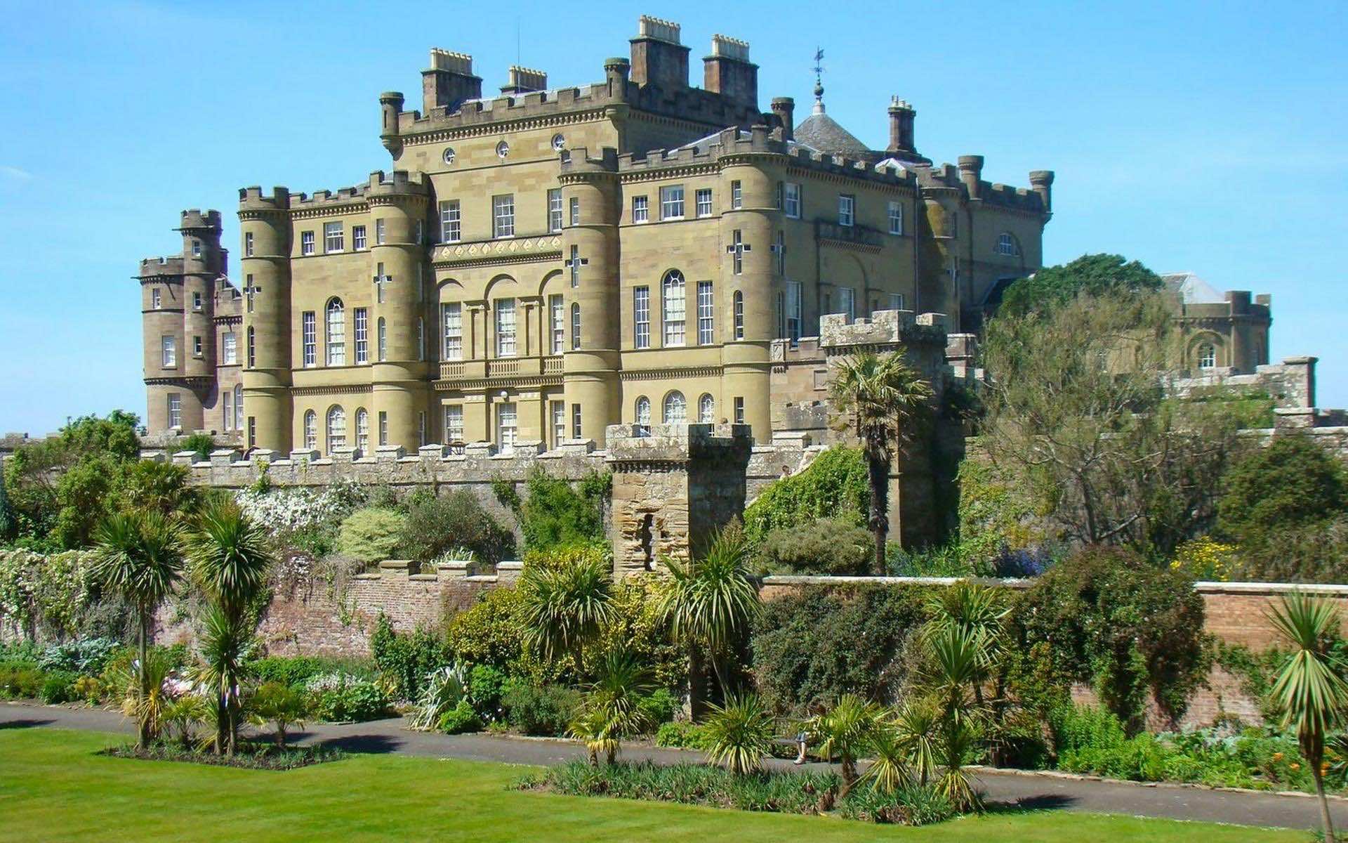A majestic castle stands proudly against a clear blue sky, surrounded by lush green gardens and a fortified wall