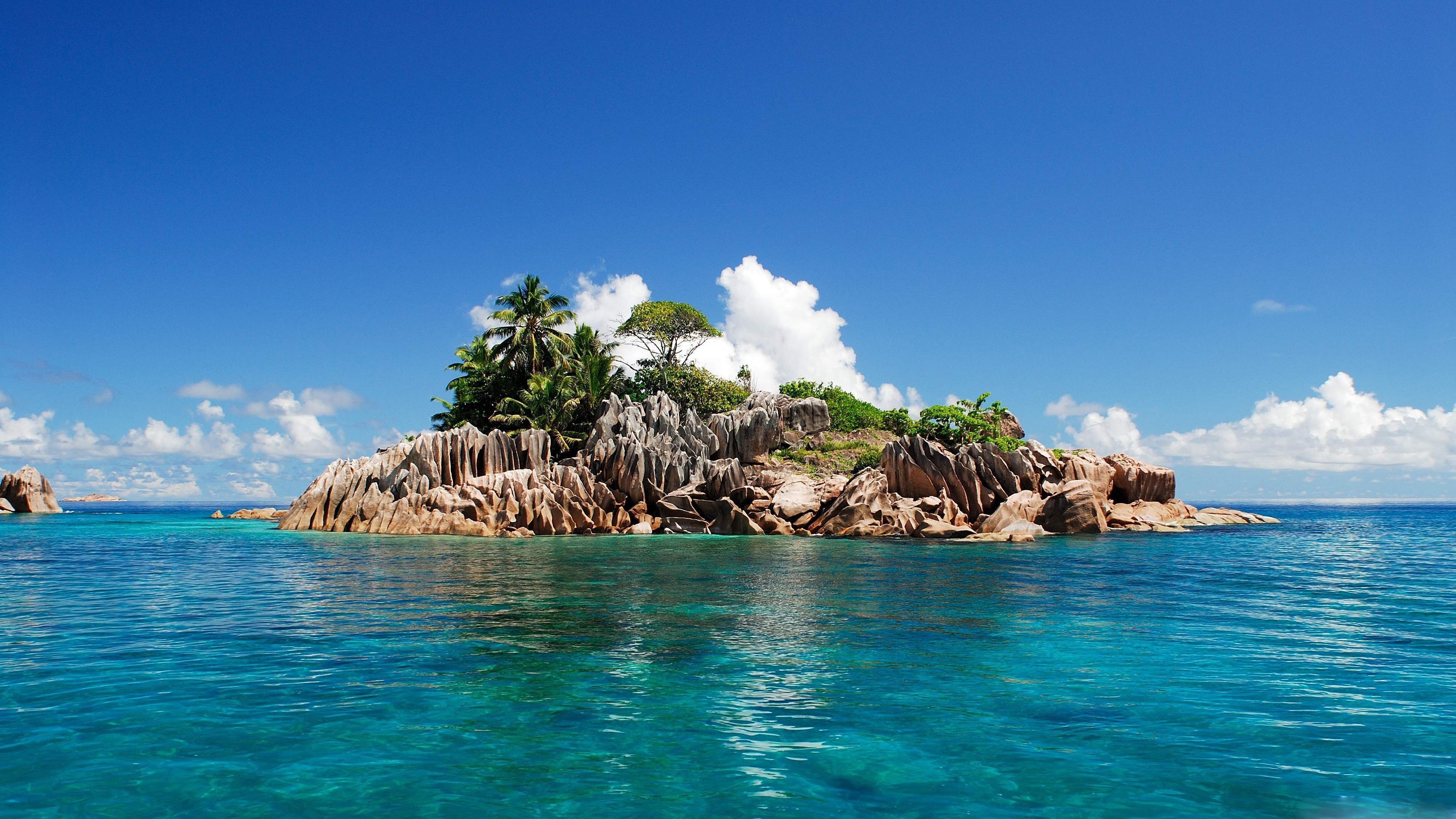 A stunning 4K HD wallpaper of a tropical island paradise with crystal clear turquoise water, granite boulders, lush palm trees, and a vibrant blue sky dotted with fluffy white clouds