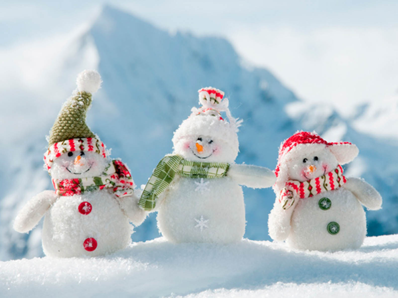 Three adorable snowmen toys standing in the snow with a snowy mountain in the background