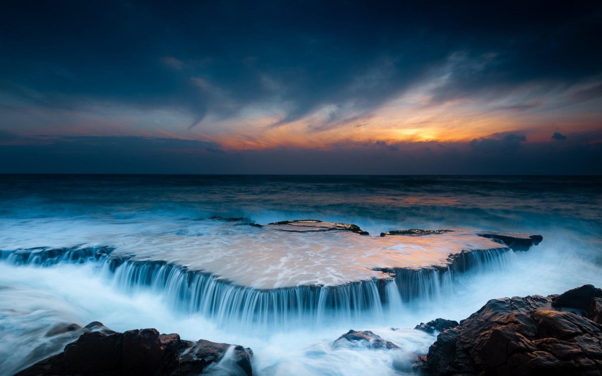 A breathtaking long exposure photograph of a waterfall-like effect created by waves flowing over a flat rock formation during a vibrant sunset at the sea
