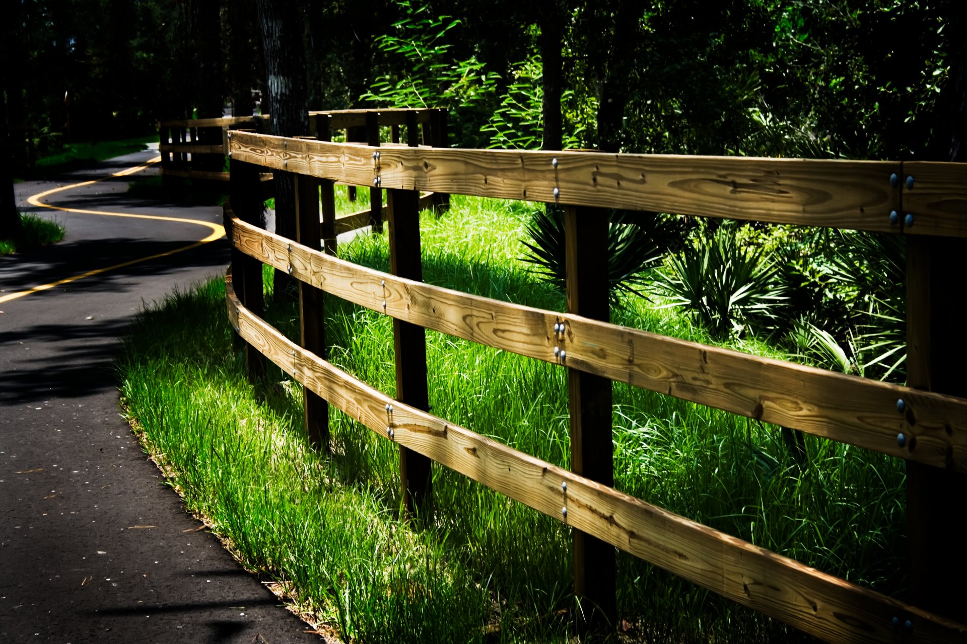 A scenic 4k wallpaper of a winding paved path bordered by a wooden fence and lush green grass, leading into a shaded wooded area