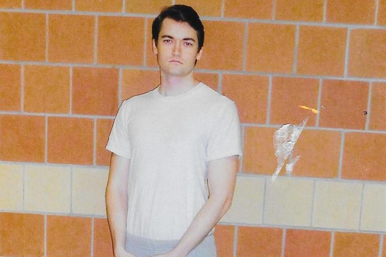 Ross Ulbricht, founder of Silk Road, in a simple white t-shirt and grey pants, stands against a brick wall