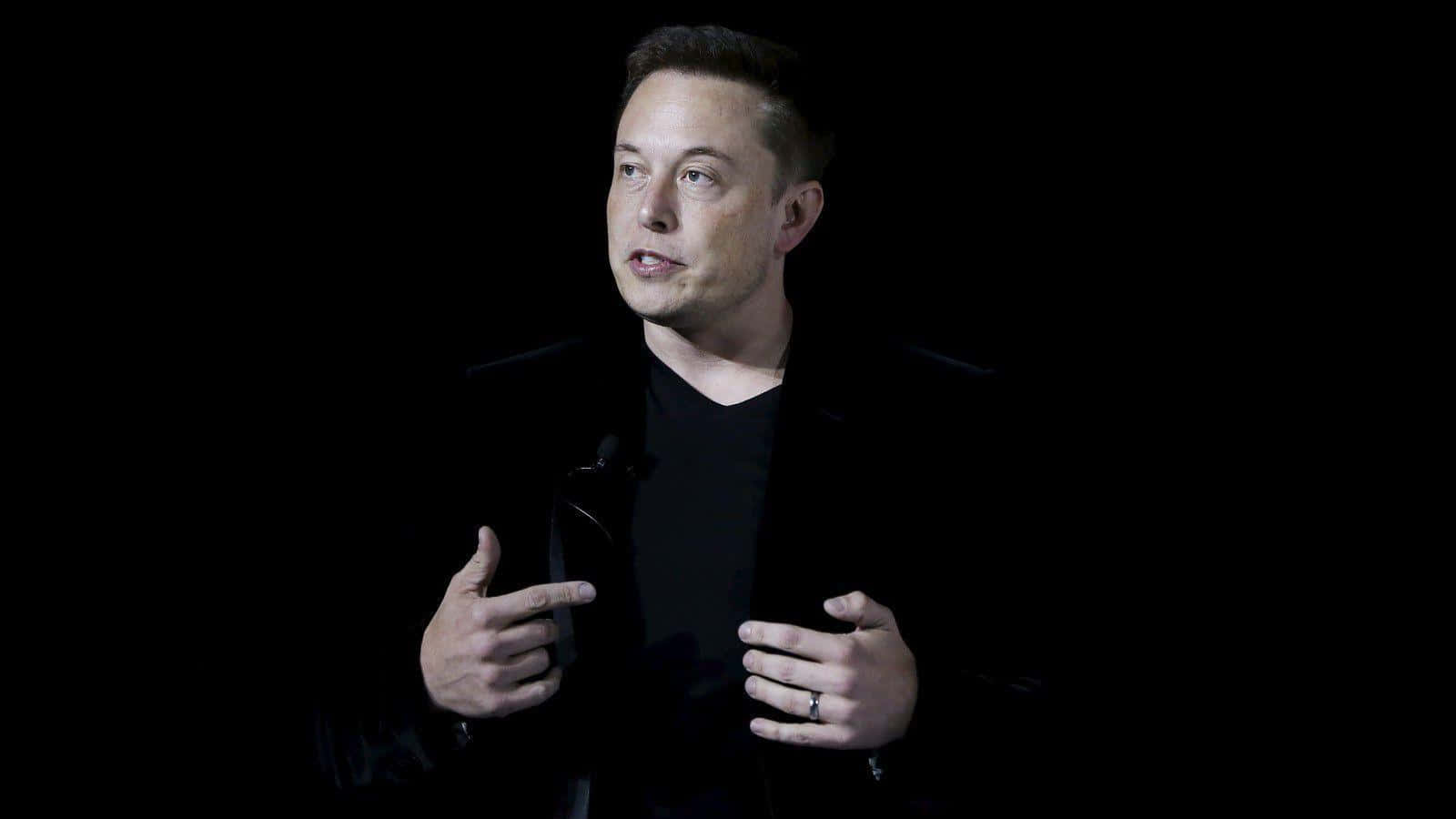 Elon Musk speaking at an event, wearing a black shirt and jacket