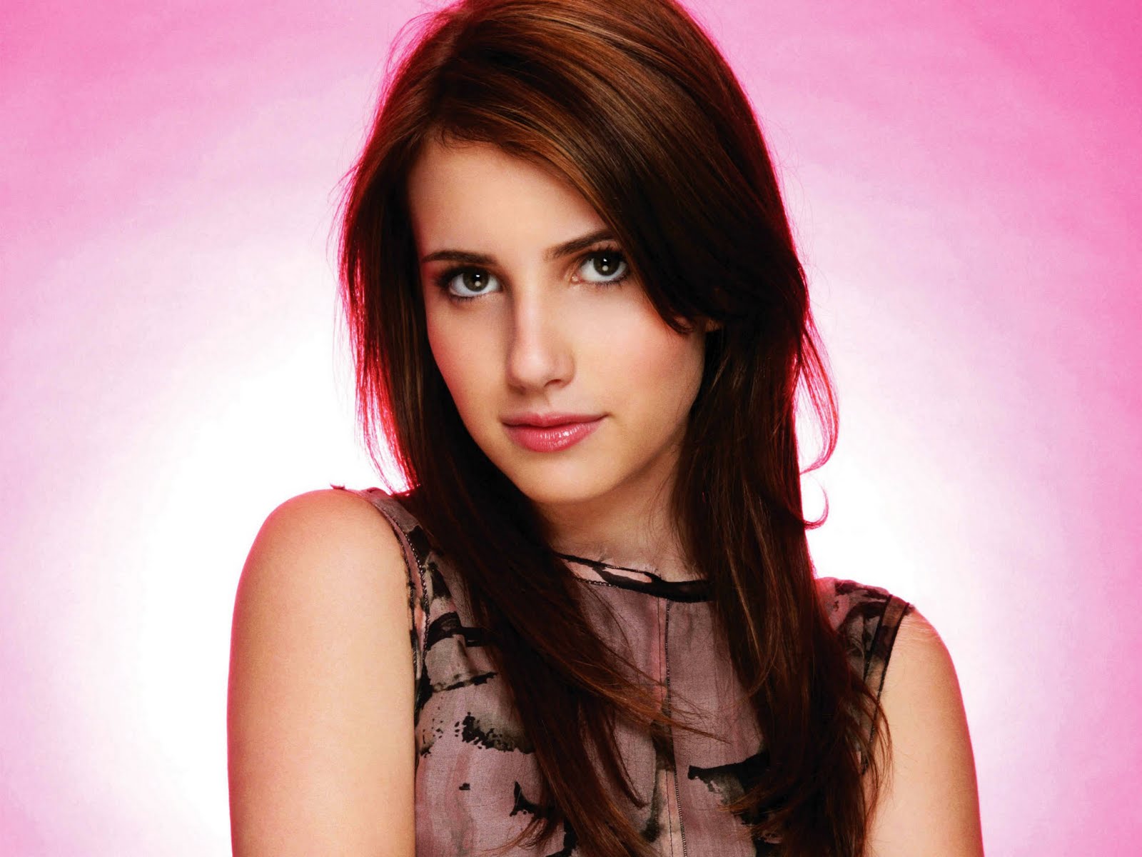 A young woman with long brown hair and brown eyes looks directly at the camera. She wears a sleeveless top, and the backdrop is a vibrant pink