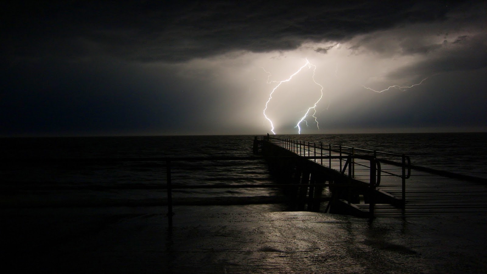 A dark and moody image of a pier stretching out into a stormy sea, lit by a dramatic lightning strike. Perfect for evoking feelings of solitude and contemplation