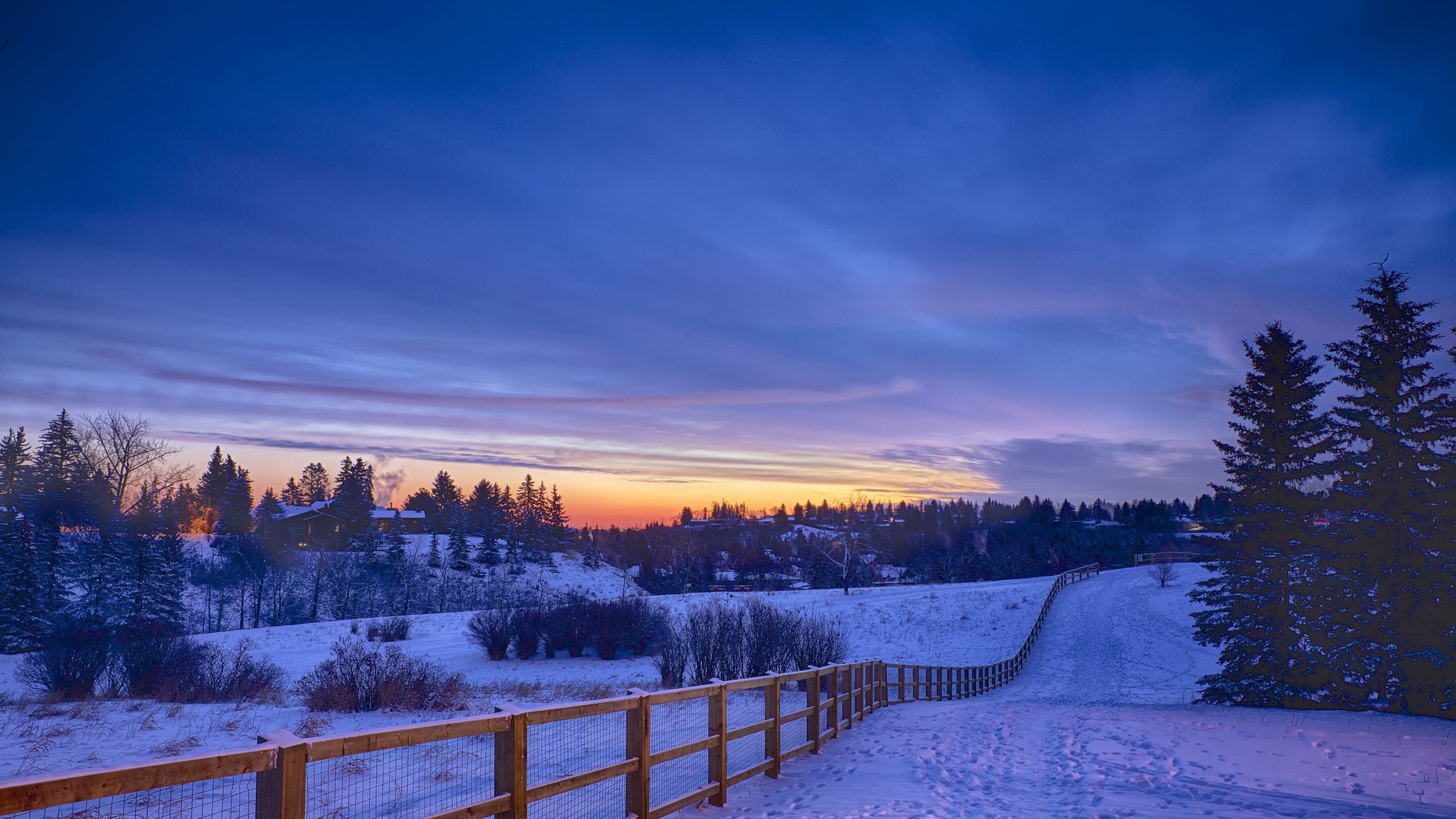 A breathtaking winter landscape at sunset, with snow-covered fields, a wooden fence, and silhouettes of trees against the colorful sky