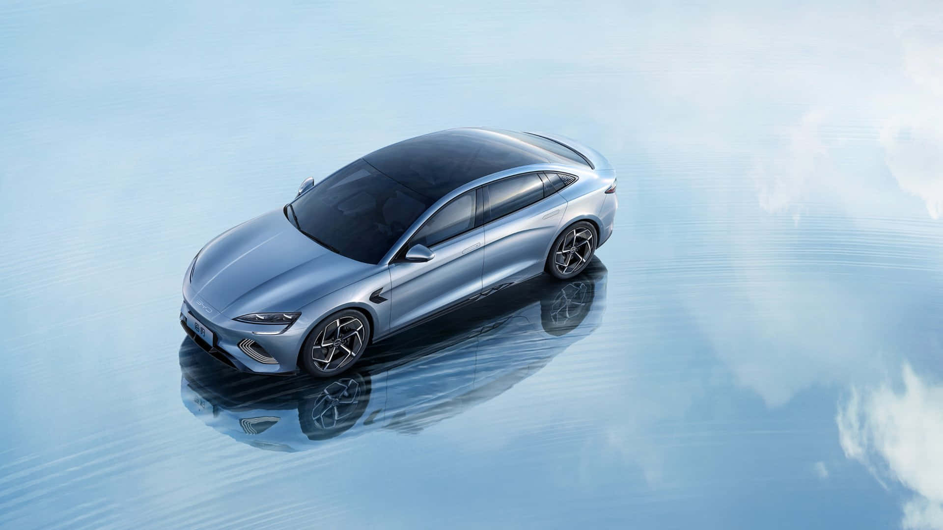 A sleek, silver BYD Seal electric sedan parked on a serene, reflective surface, possibly water, with the sky and clouds mirrored below