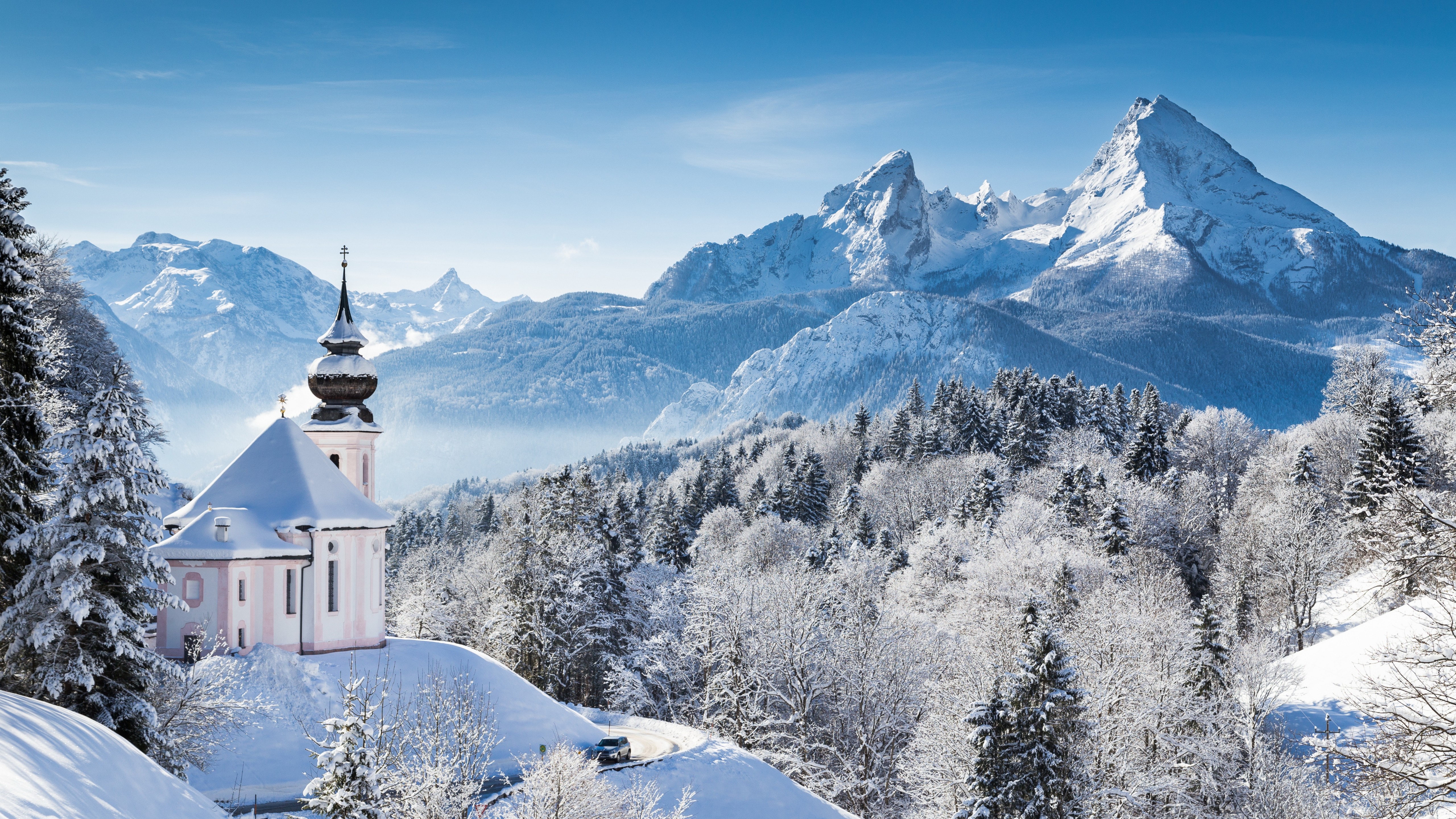 A picturesque winter landscape with a snow-covered church in the foreground and majestic, snow-capped mountains in the background