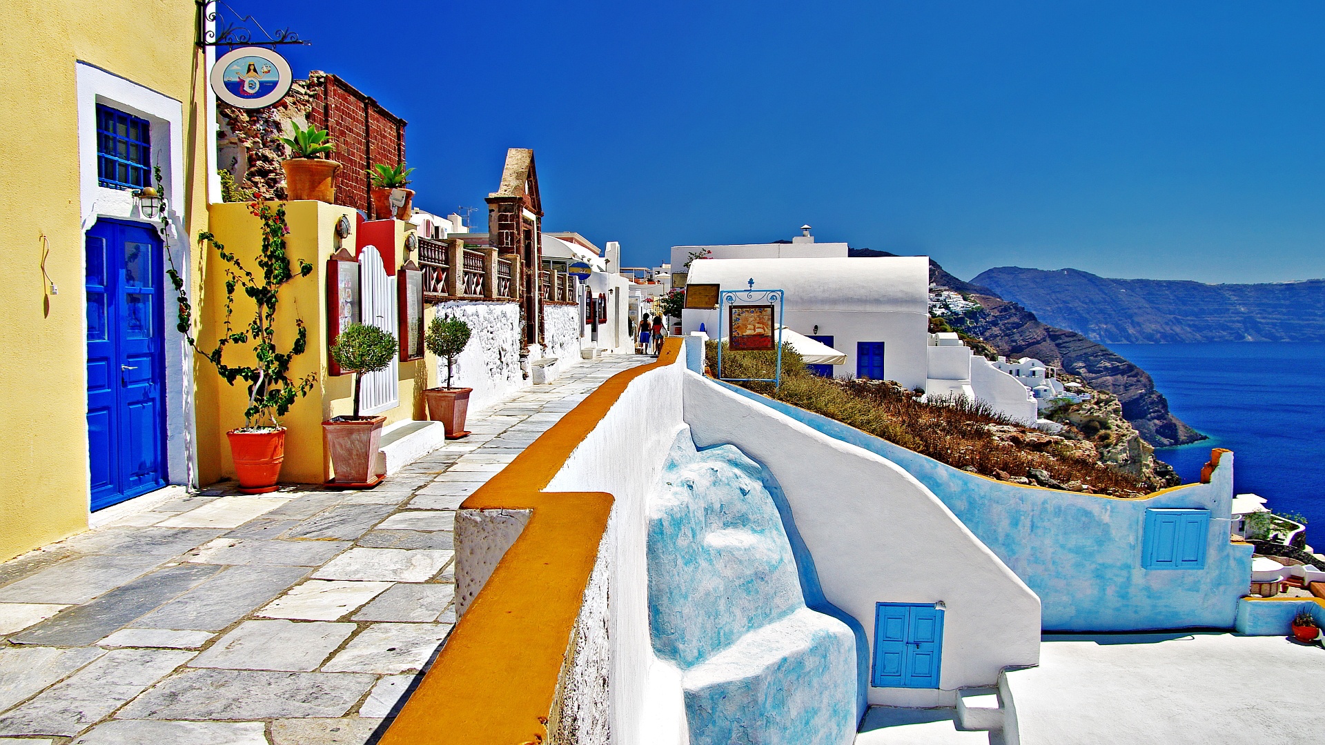 A picturesque walkway in Santorini, Greece, with whitewashed buildings and vibrant blue accents, overlooking the Aegean Sea