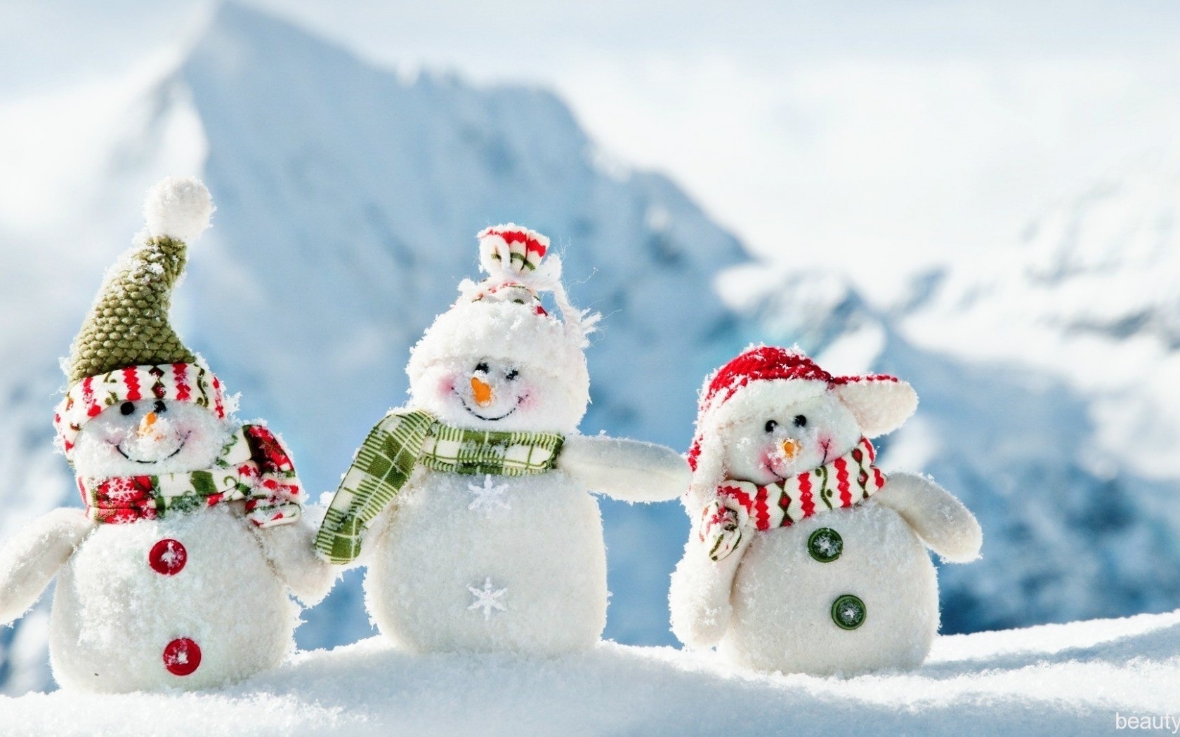 Three adorable snowmen toys stand in the snow with a backdrop of snowy mountains. They wear festive hats and scarves