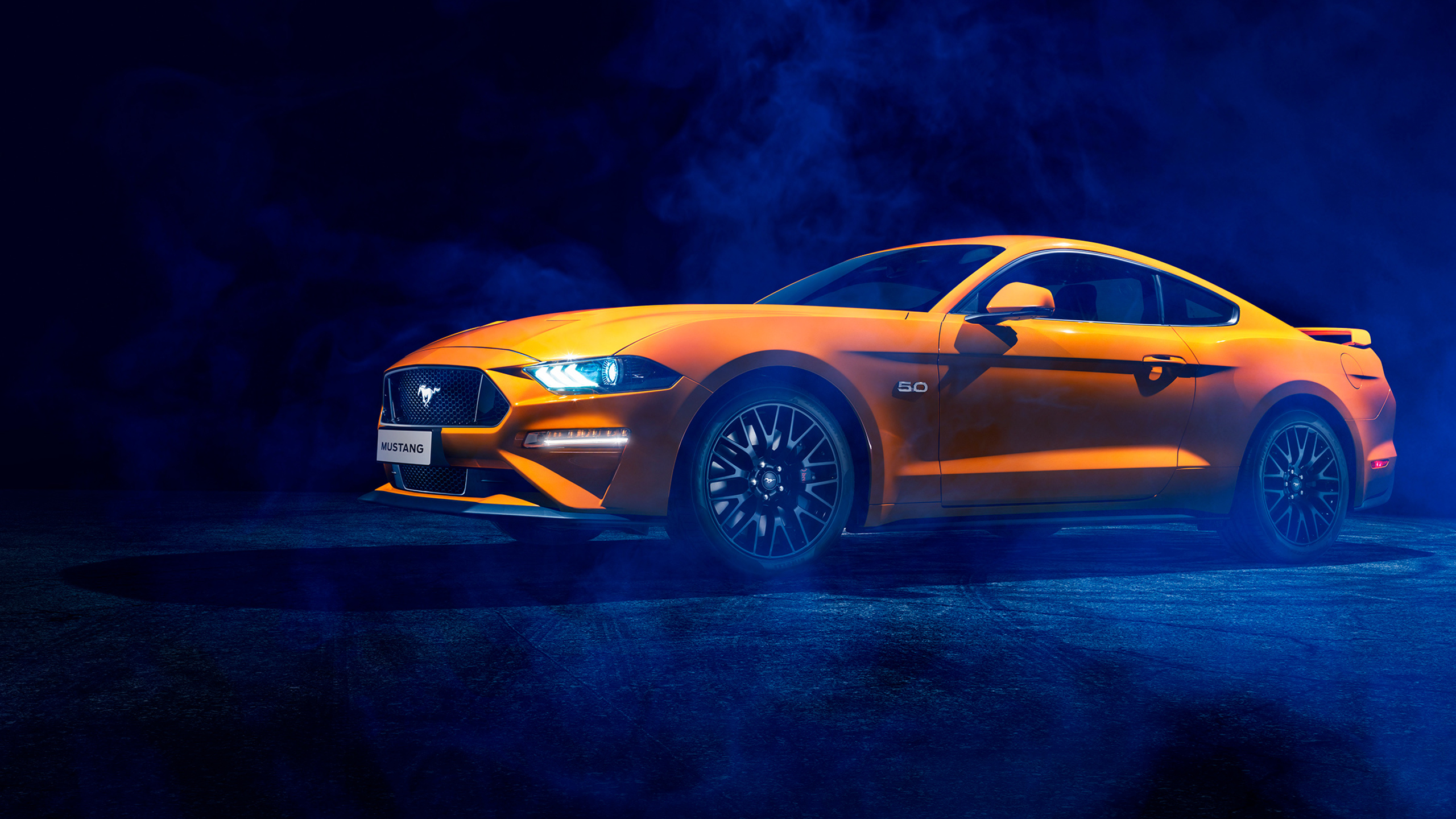 A vibrant orange Ford Mustang coupe, illuminated by dramatic lighting, against a dark, smoky backdrop