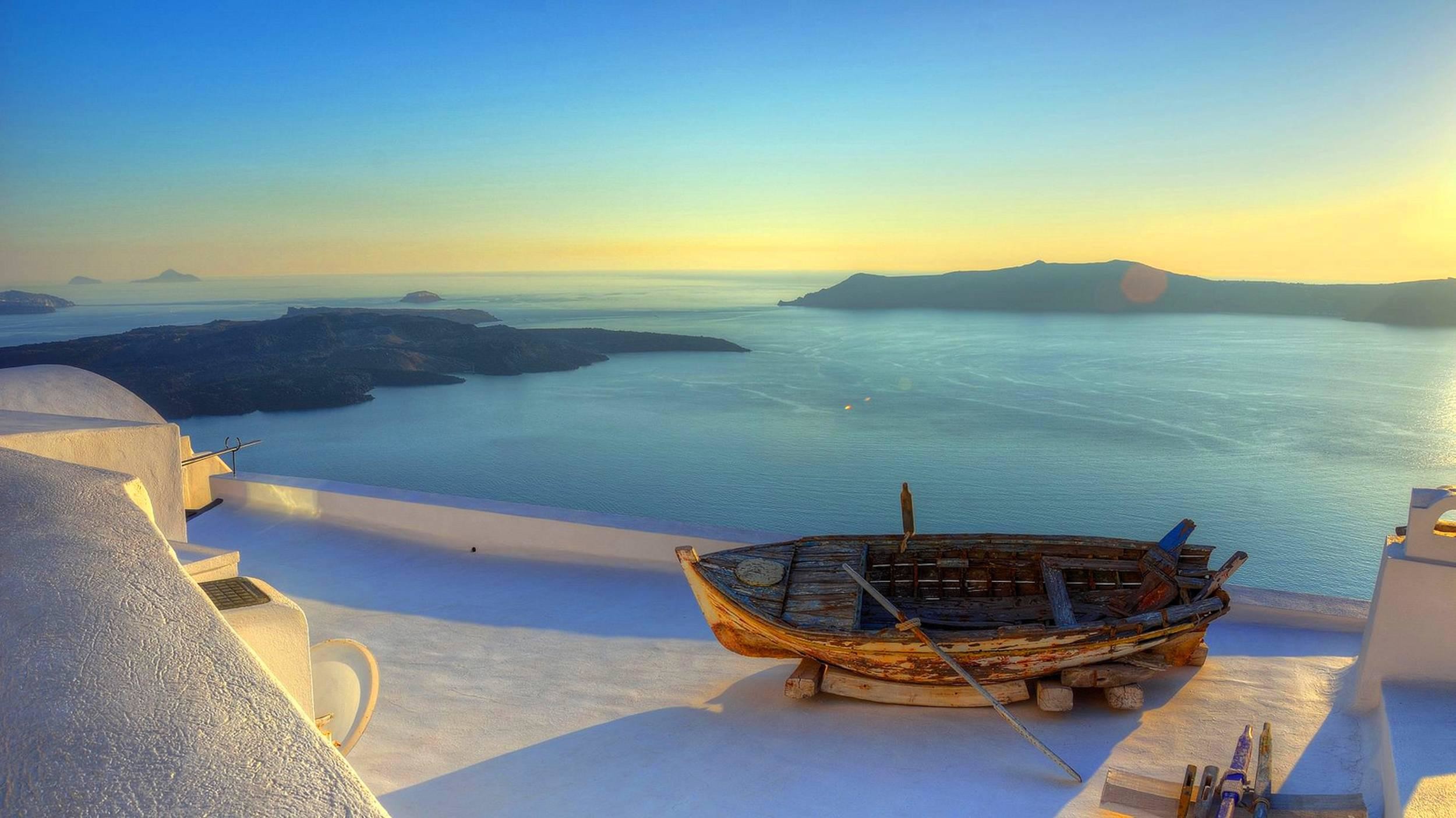 A breathtaking sunset over the caldera in Santorini, Greece. A weathered wooden boat rests on a white rooftop, overlooking the Aegean Sea and nearby islands