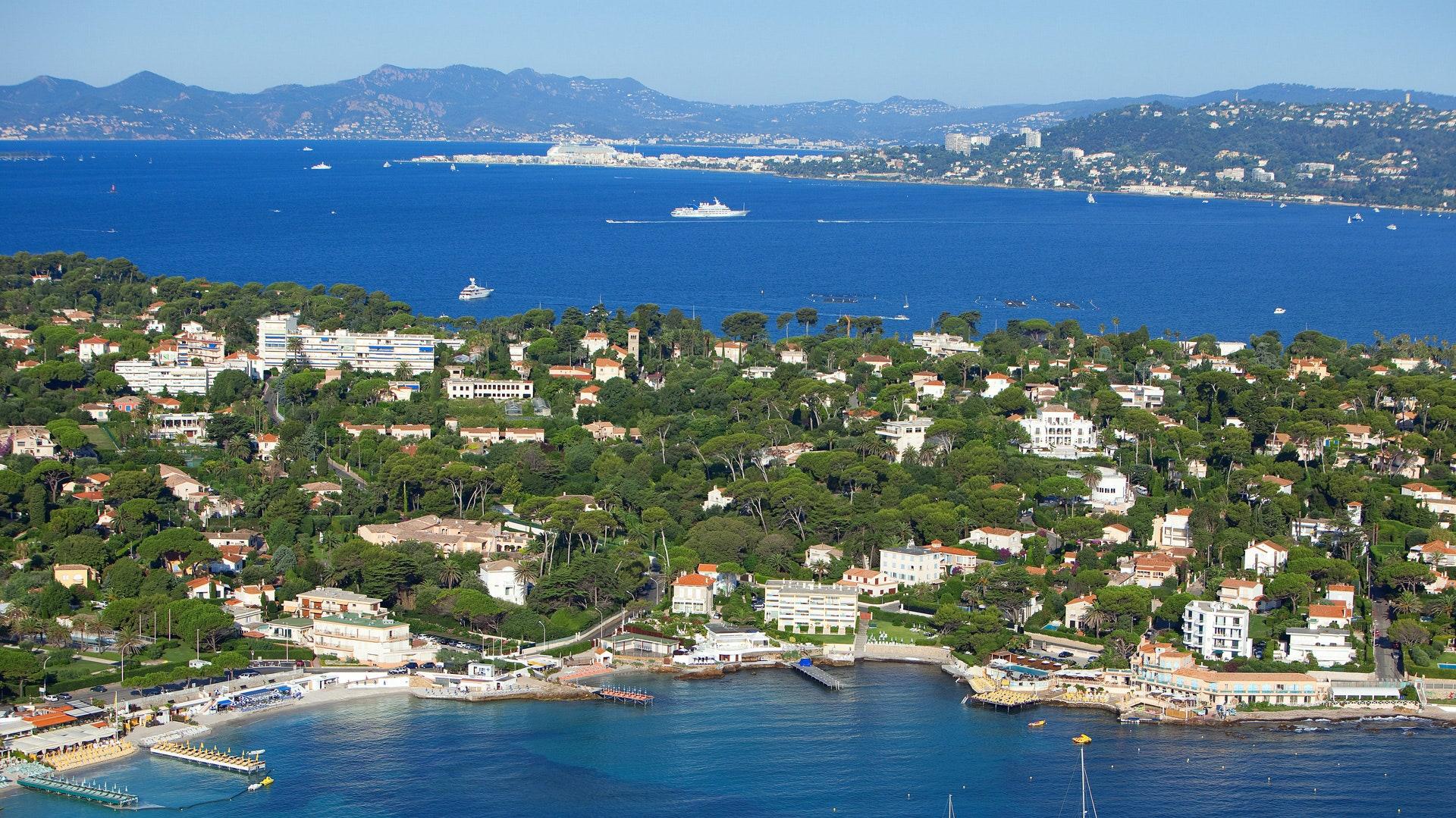 Beautiful Antibes background image featuring blue ocean with a large cruise ship sailing in the background surrounded by lush green trees and houses providing a stunning view of the coastal area