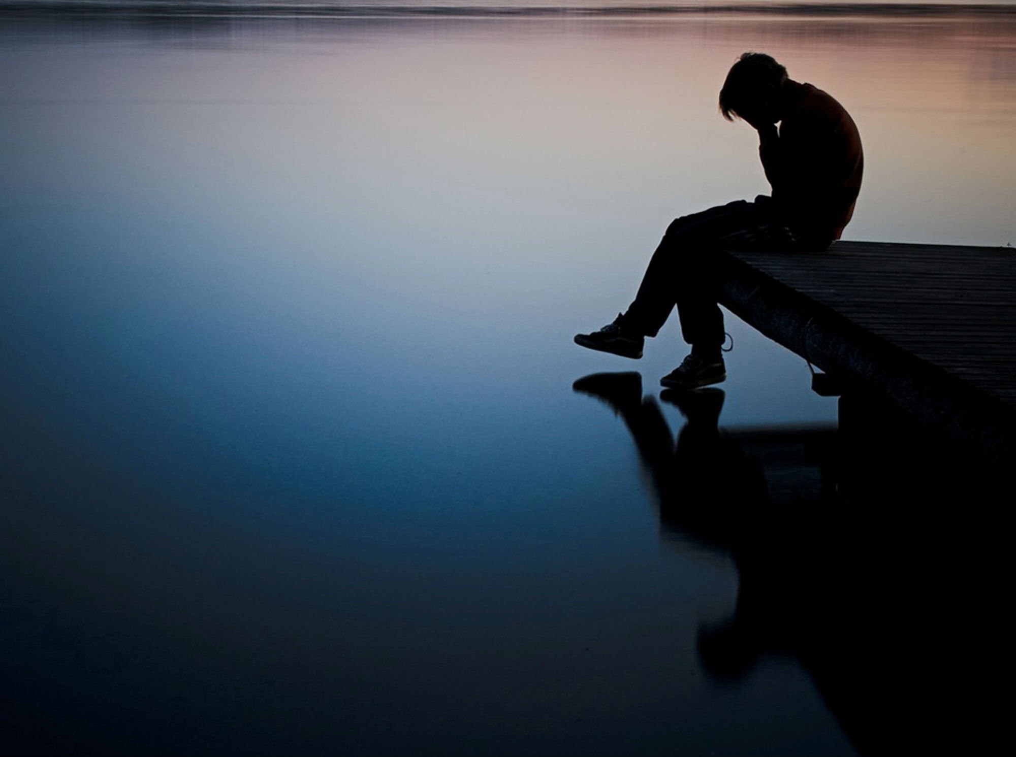 Silhouette of a lonely person sitting on a dock at sunset, reflecting on the still water, evoking a sense of sadness and contemplation