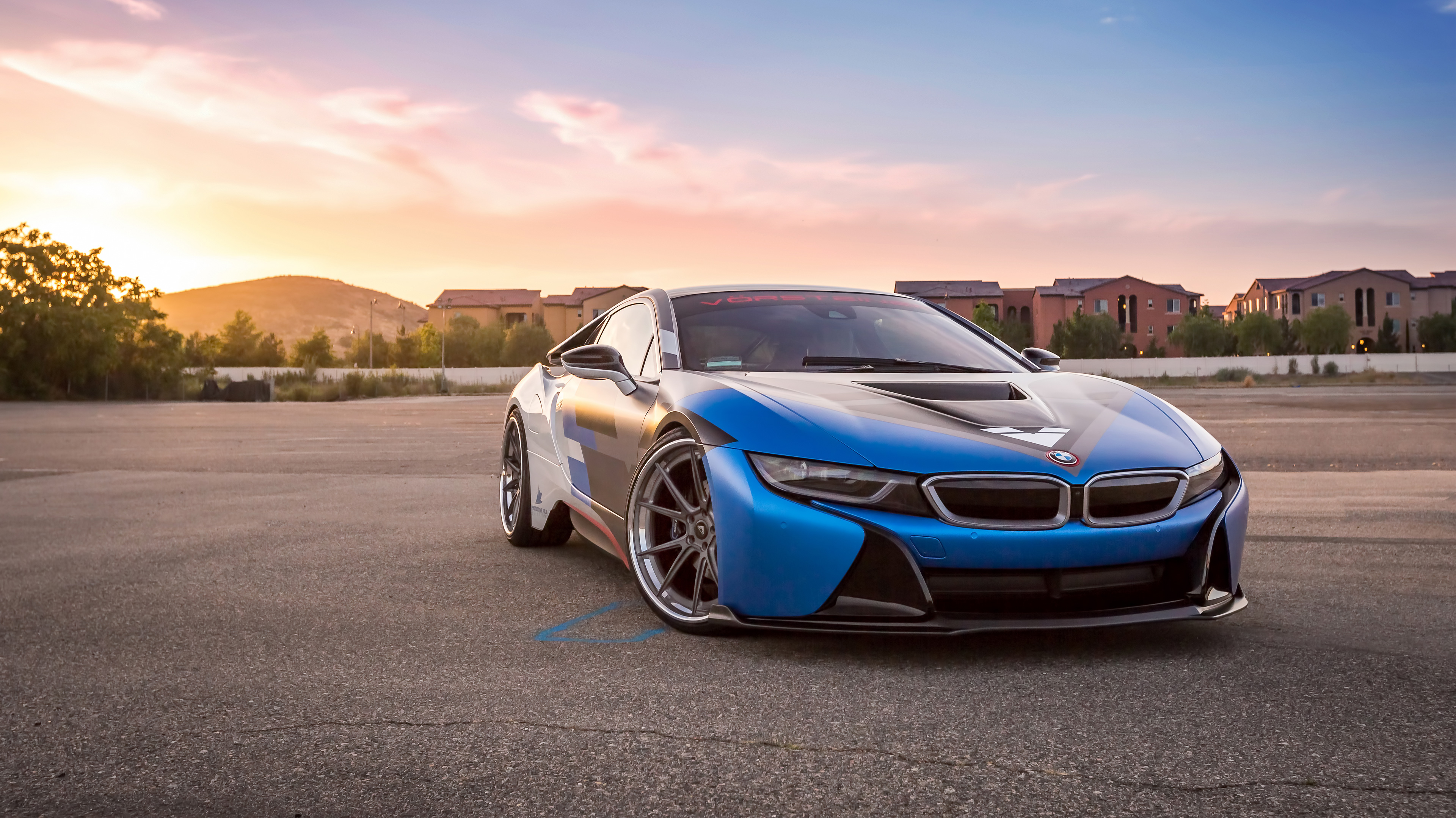 A customized BMW i8 sports car parked on a paved surface during sunset. The car is predominantly blue with black accents and features aftermarket wheels. The background includes a hillside and residential buildings