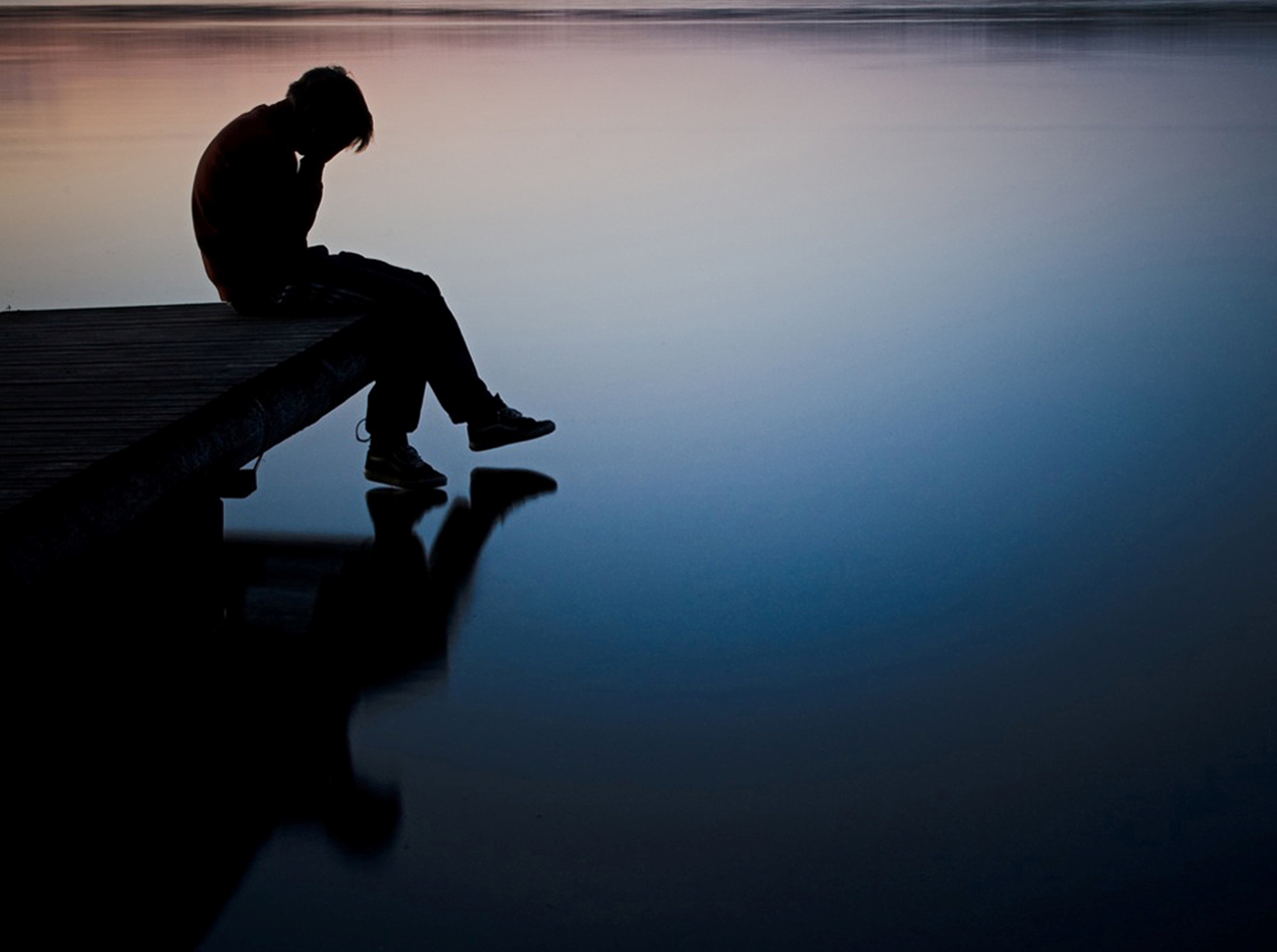Silhouette of a lonely person sitting on a dock at sunset, reflecting on the calm water, evoking a sense of sadness and contemplation
