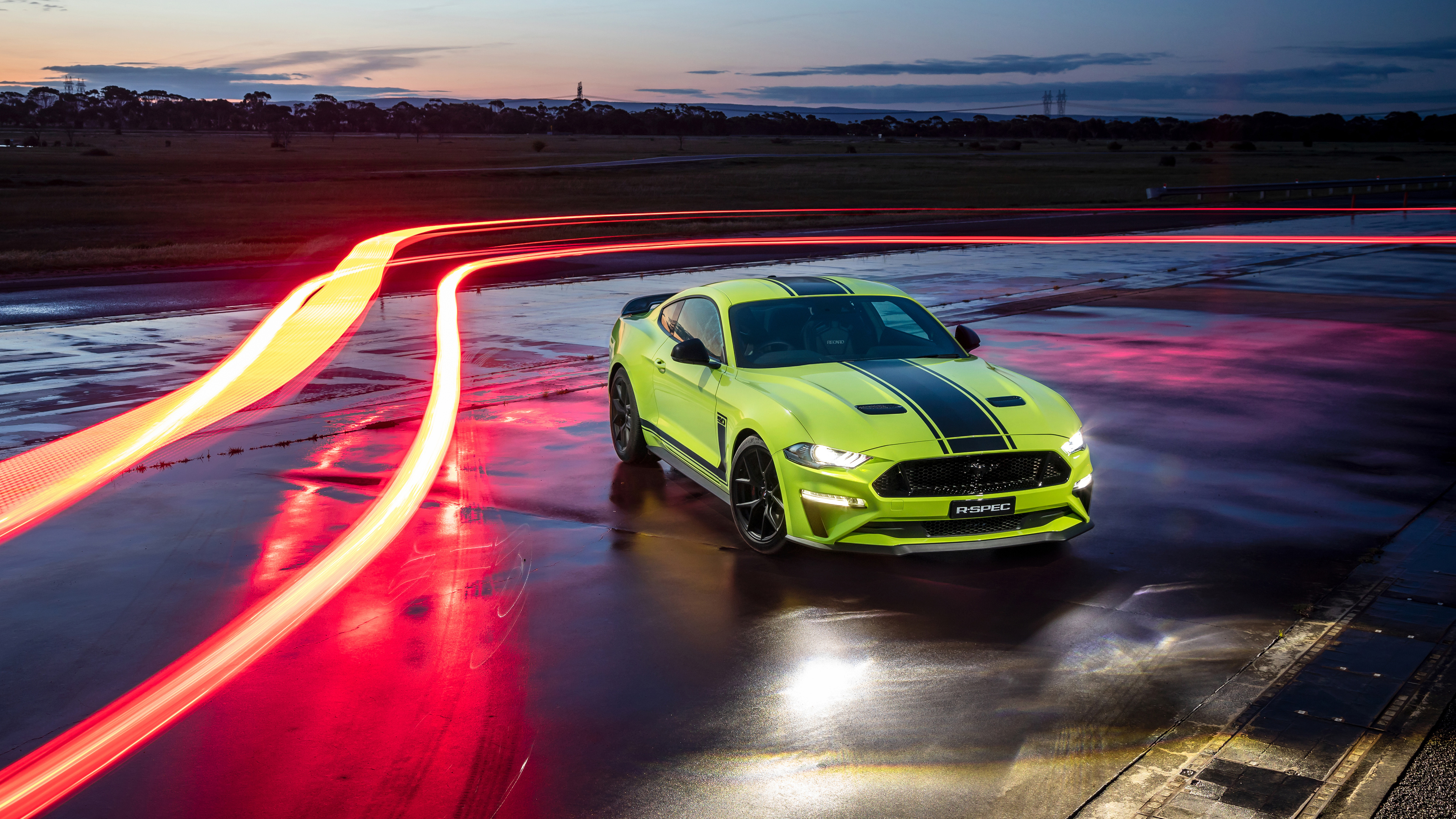 A vibrant green Ford Mustang R-Spec parked on a wet track at dusk, with light trails streaking behind it, creating a dynamic and sporty atmosphere