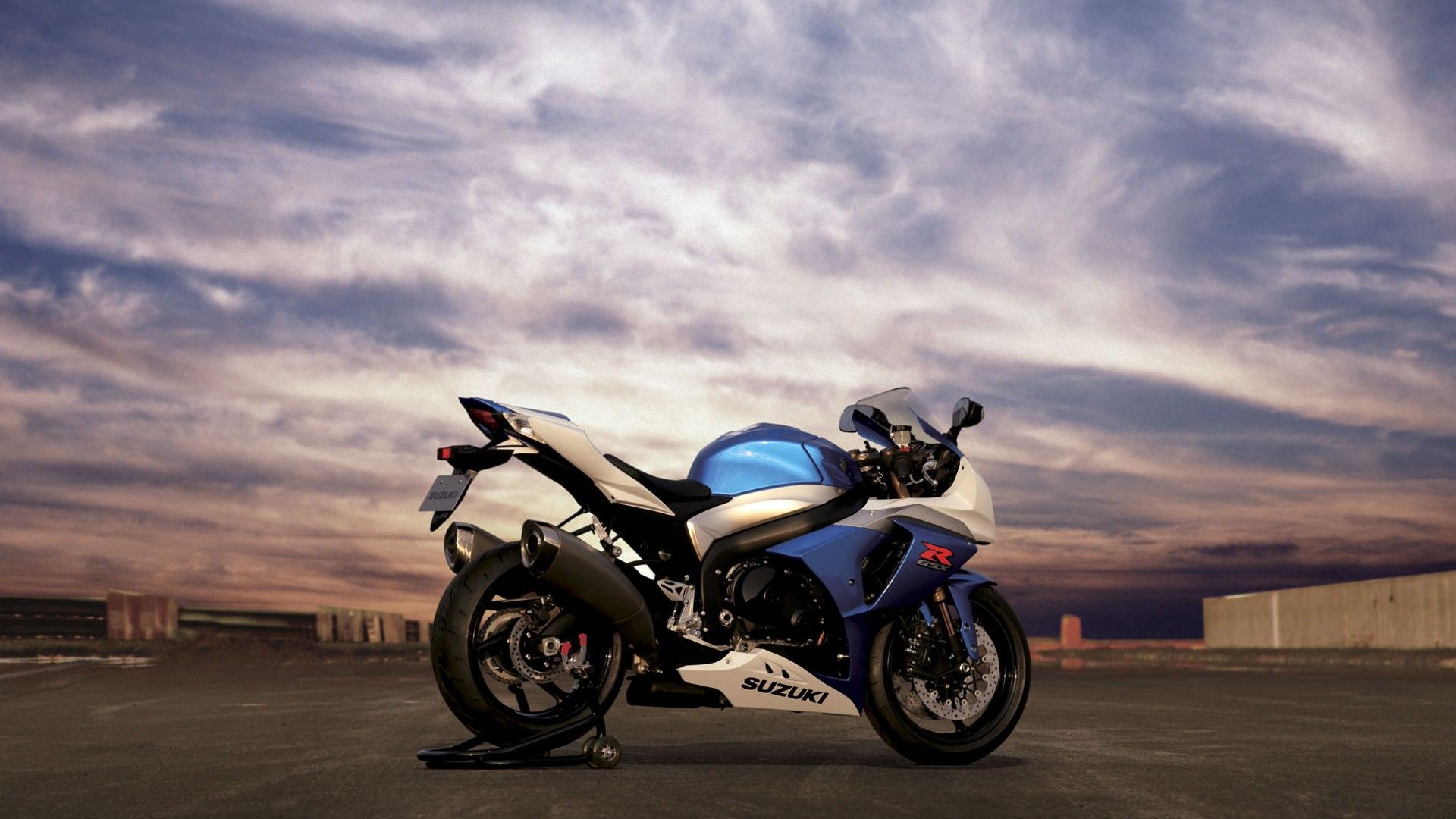 A blue and white Suzuki GSX-R sportbike parked on a paved surface against a dramatic sky backdrop