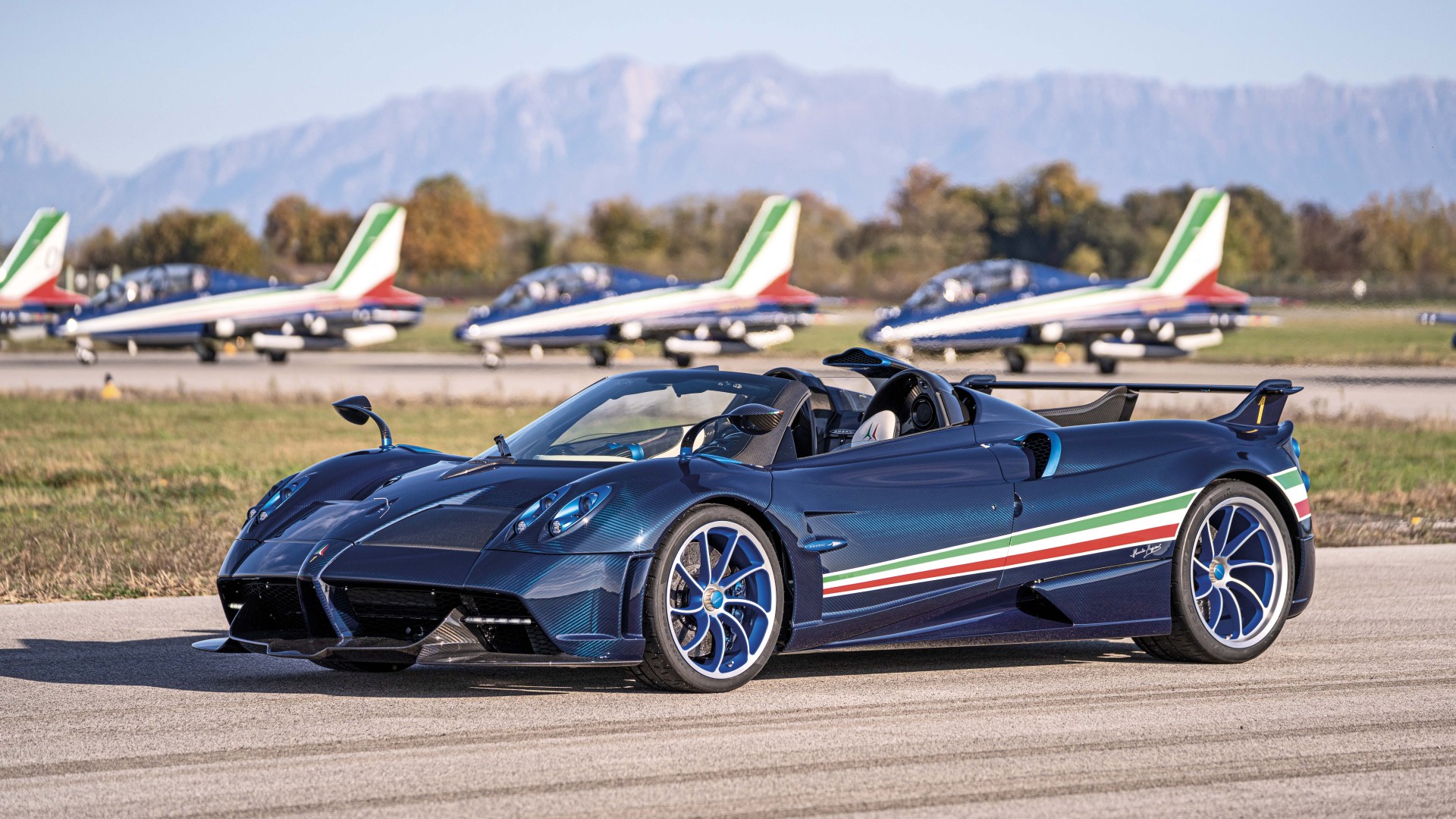 A stunning Pagani Huayra Roadster BC in exposed carbon fiber with the Italian flag colors, parked on an airfield with fighter jets in the background