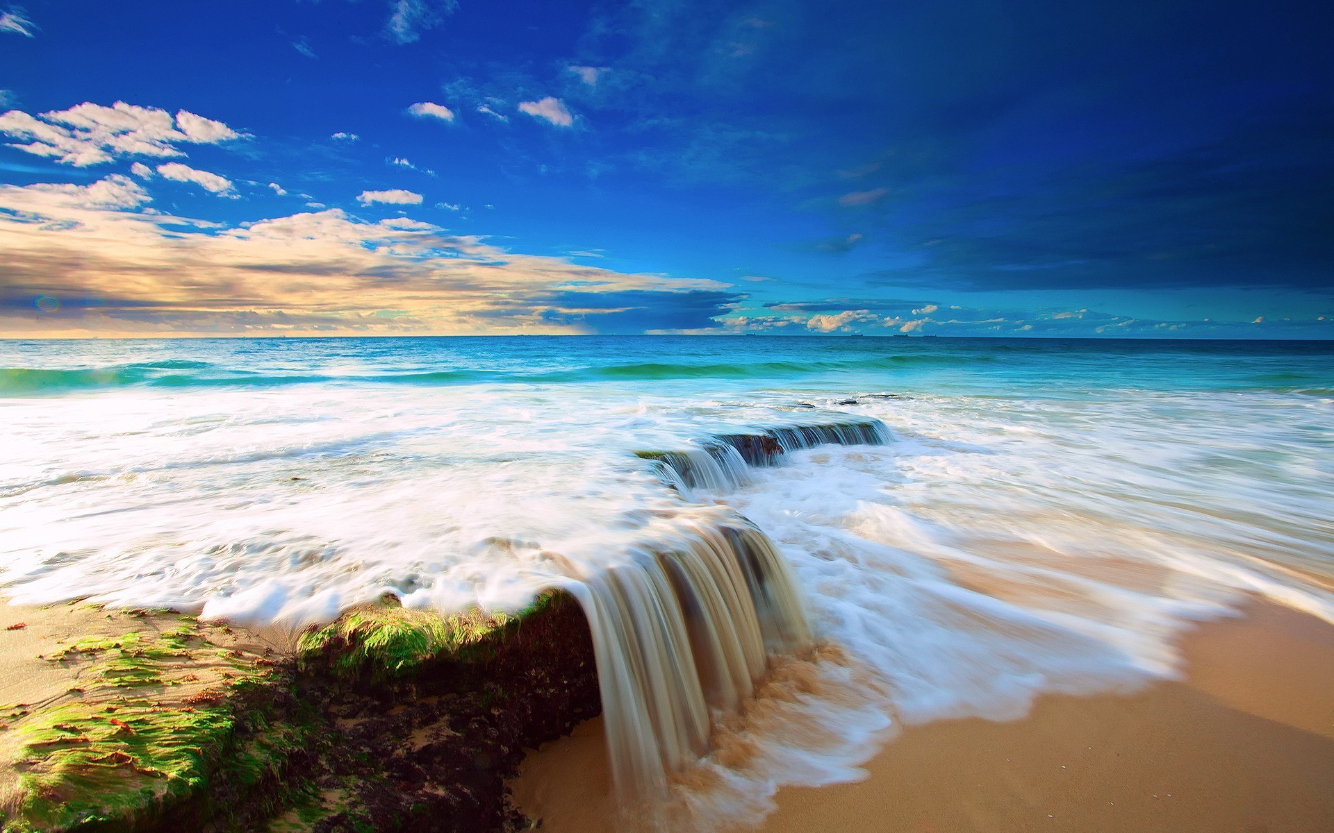 A cascading waterfall flows onto a sandy beach, with the turquoise ocean and a vibrant sunset in the background